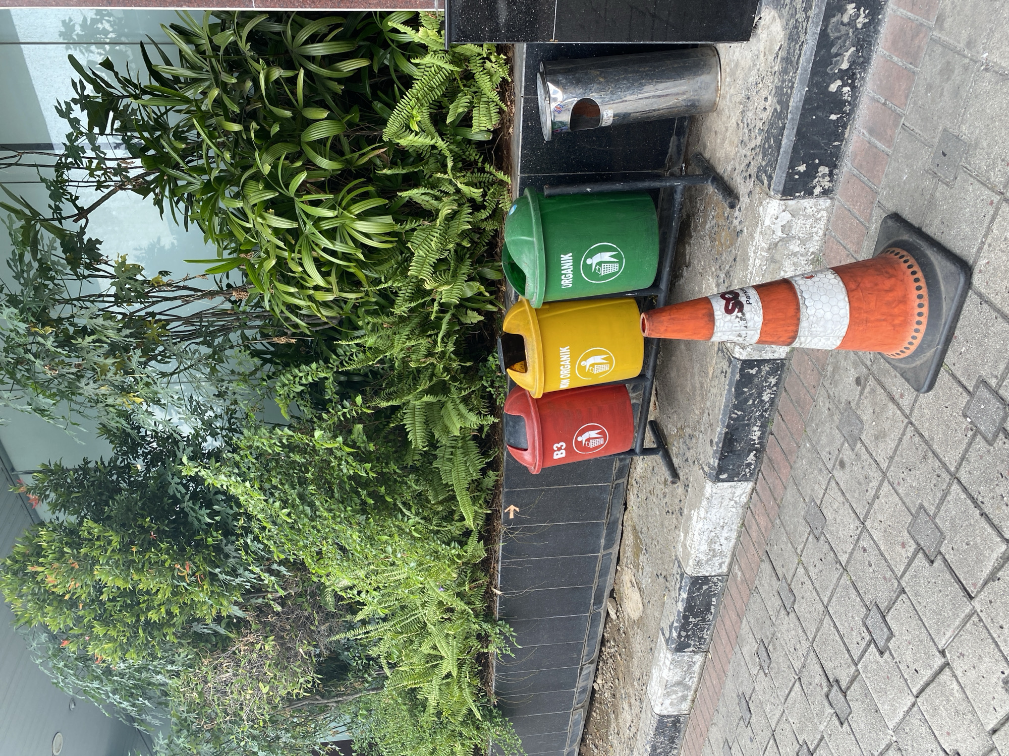 In South Jakarta, Indonesia, a set of three colored trash bins—red, yellow, and green—are positioned outdoors next to a building with lush greenery. The red bin is labeled "B3" with a pictogram of a person throwing trash into a bin. The yellow bin is marked "NON ORGANIK," also featuring a similar pictogram. The green bin is labeled "ORGANIK" and displays the same trash-throwing pictogram. To the right of these bins, a silver cylindrical trash receptacle with an open slot at the top is visible. In the foreground, an orange and white traffic cone stands on a paved walkway constructed with interlocking concrete blocks. The walkway is bordered by a curb with black and white painted markings. The background features a dense arrangement of various green plants and trees.