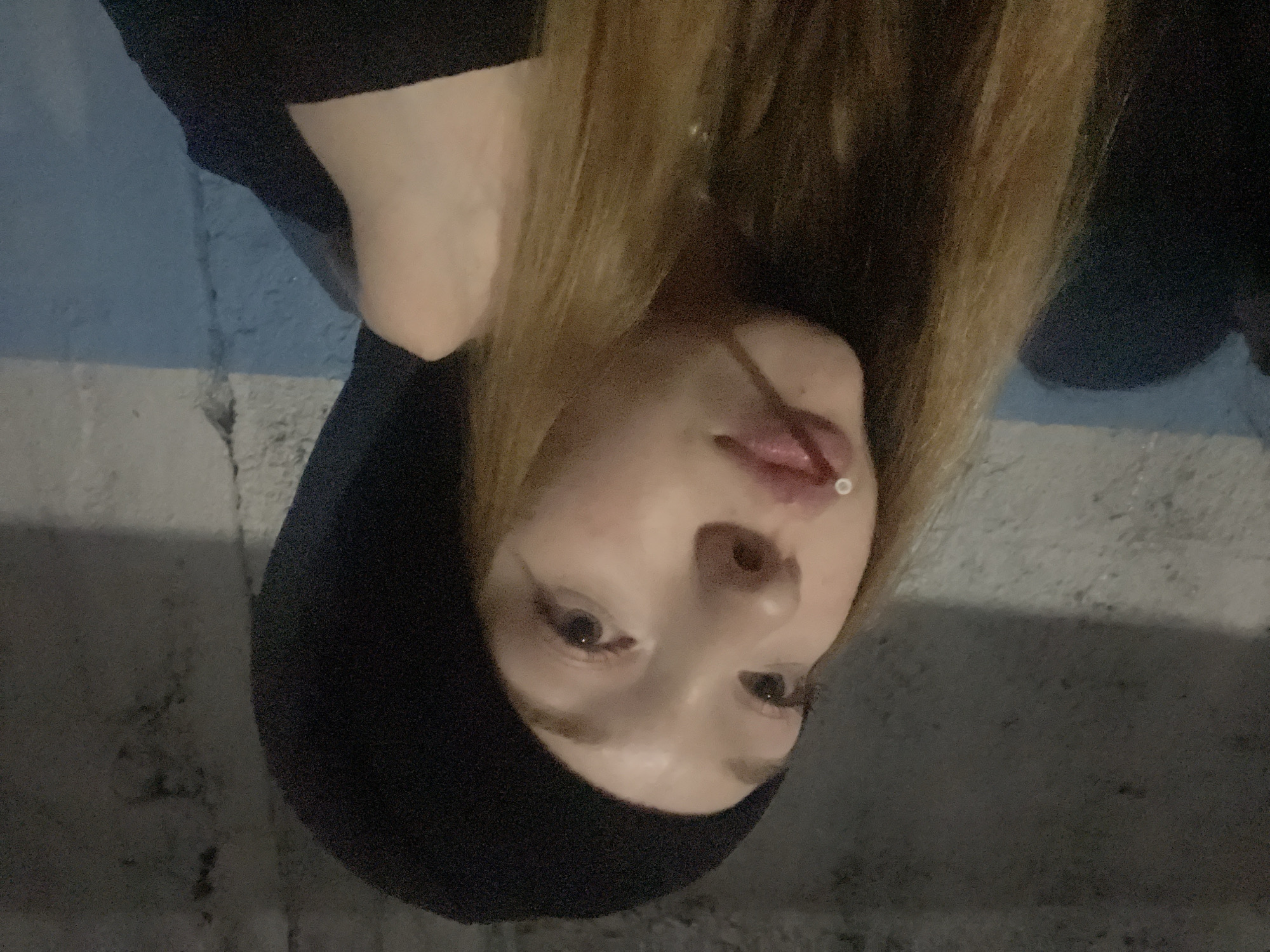 A young woman with light brown hair is shown in a close-up shot, rotated counter-clockwise. She has fair skin, brown eyes, and a lip piercing. She is wearing a black hoodie. The background appears to be an exterior wall with a blue painted section and a rough concrete surface. The location is identified as the city of Sosnovo, Russia.