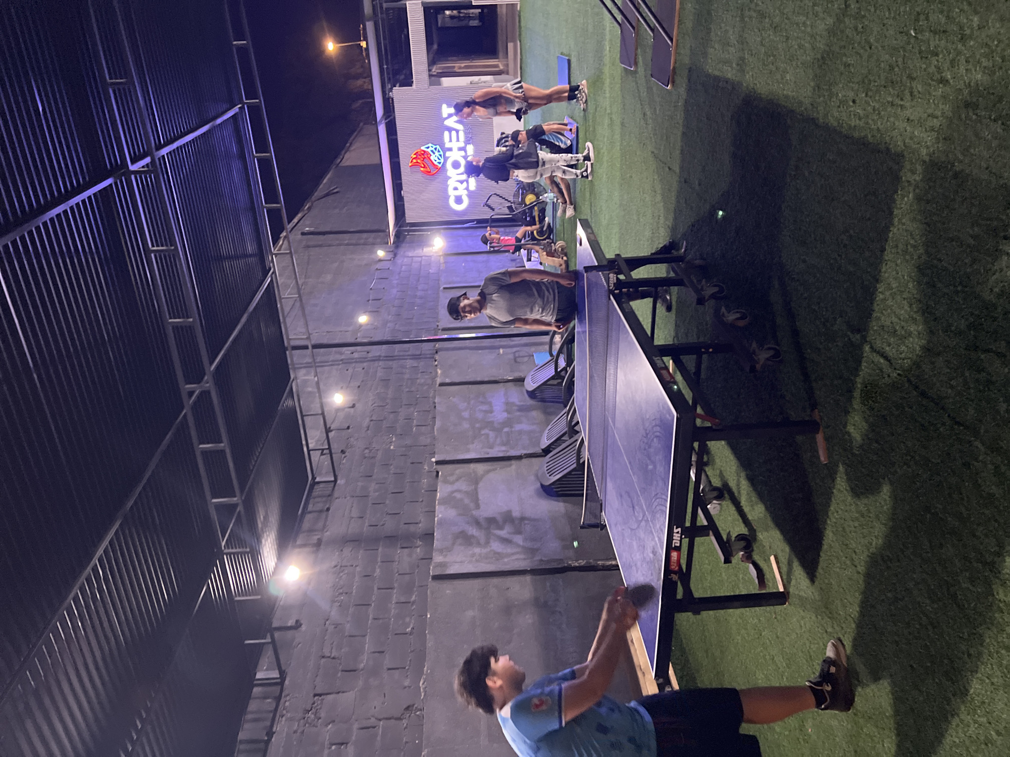 A man in a blue t-shirt is playing ping pong on a blue table outdoors. He is located in Arica, Chile. In the background, people are exercising near a building with the word "CRYOHEAT" lit up in neon letters. The environment includes an artificial grass surface, metal structures, and a brick wall.