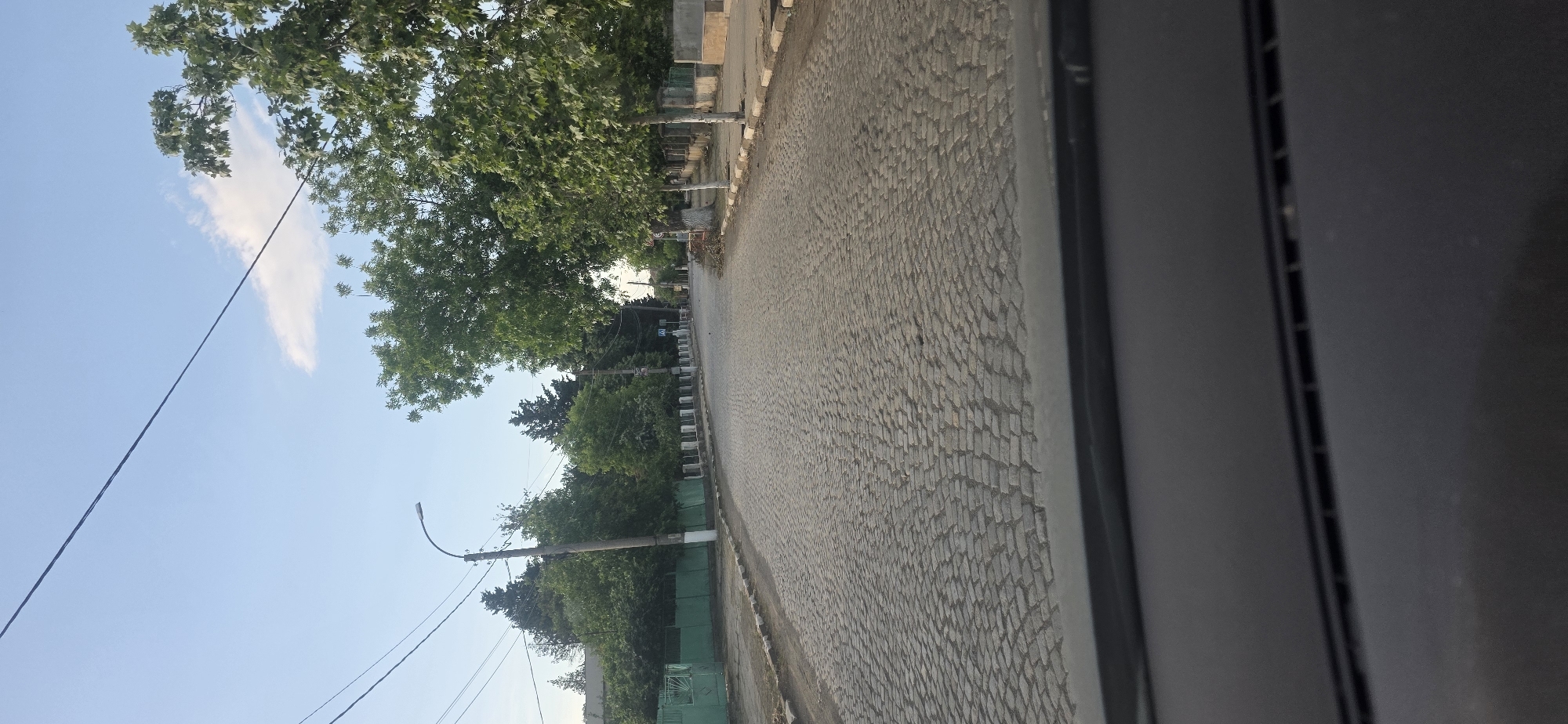 The image shows a street in Troyanovo, Bulgaria. The street is paved with cobblestones and lined with trees. A street light is visible on the left side of the road. The sky is blue with a few clouds. The photo seems to have been taken from within a building, as a dark, rectangular object obstructs part of the view on the right.