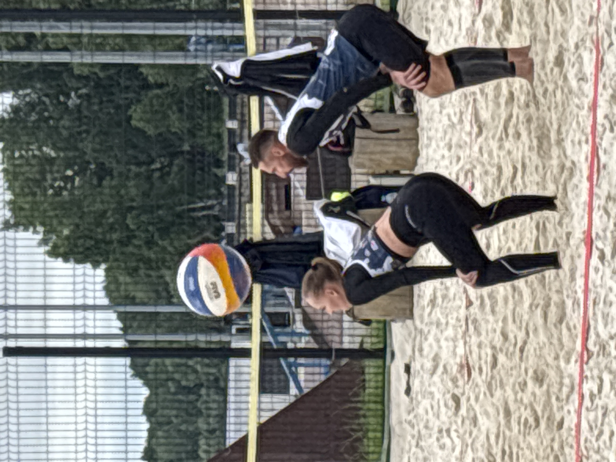 The image shows a beach volleyball game in progress at the beach volleyball courts located at the stadium in Chaikovsky, Russia. Two players are visible on the sand court, one male and one female. The female player is in the foreground, on the left, in a crouched defensive position, facing away from the camera and towards the net. She is wearing a black long-sleeved top with a patterned front and black leggings. The male player is behind her and to the right, also in a crouched position, facing the net. He is wearing a black long-sleeved shirt with blue and white accents on the front and black shorts, with black compression sleeves on his calves. A multi-colored volleyball is in the air above and to the left of the players. The background shows a chain-link fence, trees, and bleachers, indicative of a sports facility. The court is made of sand, with a red boundary line visible in the foreground. The sky is overcast.