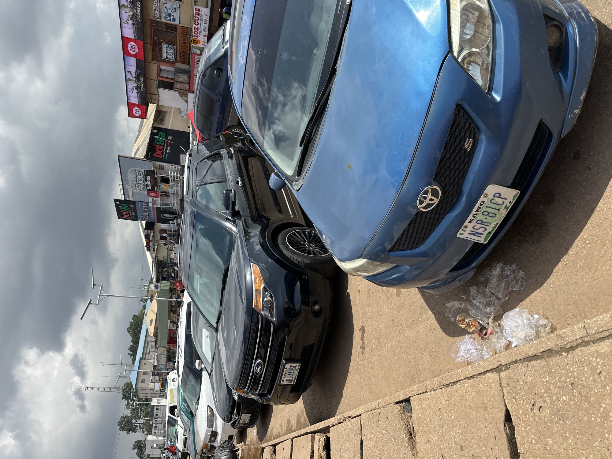 This is a photograph of a street scene in Jos, Nigeria, with several cars parked along the side of the road. The image is oriented vertically. The foreground is dominated by the front of a blue car, with a license plate that reads "NSR-81CP KANO". To the left of the blue car is a black SUV, its license plate reading "ABJ-440SC". Further to the left, other vehicles, including a white car, are visible, suggesting a parking area or a busy street.
Behind the cars, buildings line the street. Some of these buildings have large billboards and signs, indicating commercial activity. Visible text includes "itel POWER 80", "bet9ja", "SAIBAS", "DICE CUBE COMMUNICATIONS", and "Wah". The presence of these signs suggests a commercial hub or market area.
The sky is overcast with dark clouds, indicating potentially rainy or stormy weather. The lighting suggests it is daytime, but the cloud cover diffuses the sunlight. The ground is paved with concrete and there are some concrete barriers. Some scattered plastic waste is visible on the ground near the blue car. The overall impression is that of a typical urban street scene in Nigeria, with parked vehicles, commercial signage, and varied weather conditions. No people are clearly visible in the photograph.