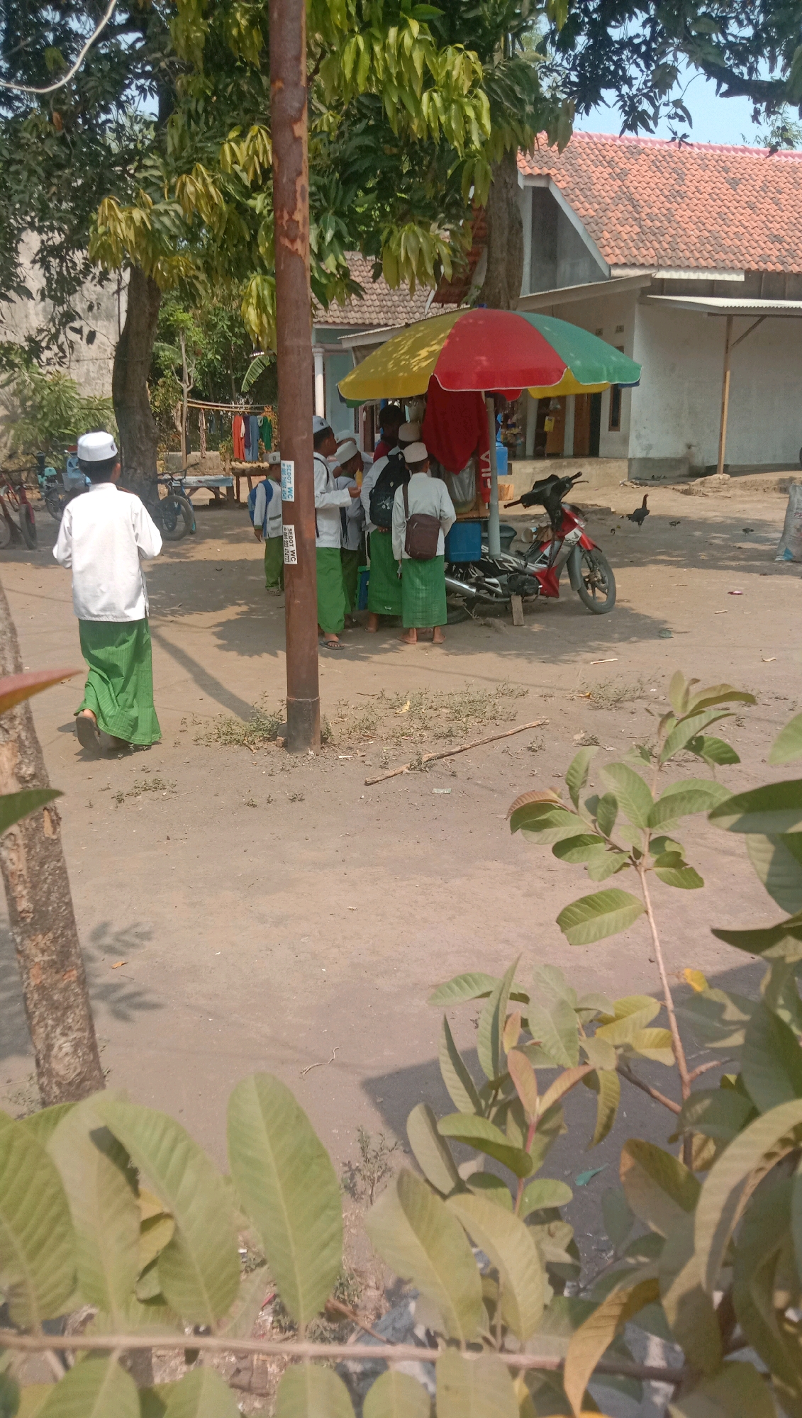 In Ngempit, Indonesia, a group of young people, likely students, are gathered around a colorful umbrella stand. The students, predominantly boys, are wearing white shirts and green sarongs with white skullcaps. Some are carrying backpacks. A man in similar attire stands to the left of the group, facing away from the camera. A red and white motorcycle is parked near the umbrella stand. The environment appears to be a rural or suburban area with dirt ground, scattered trees, and houses in the background with terracotta tile roofs. Clothes hang on a clothesline behind the students, and a chicken can be seen in the background. The scene is lit by bright sunlight, casting shadows.