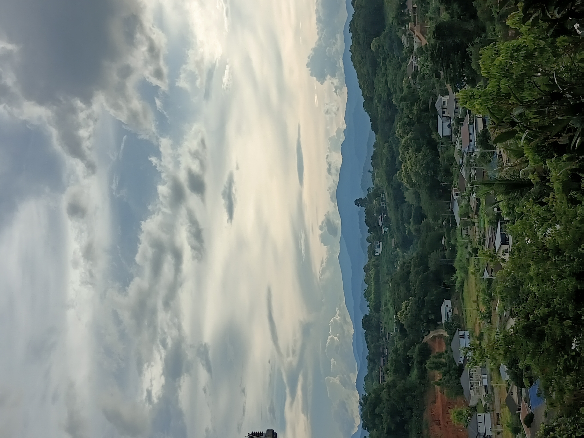 The image depicts a panoramic view of a rural residential area nestled within a lush, hilly landscape, likely in or near บ้านเหล่าพัฒนา, Thailand. The foreground is dominated by dense green foliage, with the tops of trees and shrubs obscuring the lower portions of the scene.
Beyond the immediate vegetation, the midground reveals a scattering of houses, varying in architectural style and color. Some houses are multi-story, with visible windows and roofs. These residences are interspersed with patches of cleared land, likely gardens or open spaces, and winding dirt paths. The terrain is uneven, with slopes and hills characteristic of the region.
The background features a series of rolling hills covered in dense forest, stretching towards the horizon. In the far distance, a range of larger mountains is visible, shrouded in a light blue haze, suggesting atmospheric perspective. The sky above is a dramatic expanse of clouds, with a mix of dark, heavy formations and lighter, brighter areas where sunlight breaks through. The lighting suggests it might be late afternoon or early evening, with the sun casting a soft glow. On the far left of the image, a portion of a tall, dark structure, possibly a utility pole or antenna, is visible against the sky. The scene is devoid of any visible people or animals.