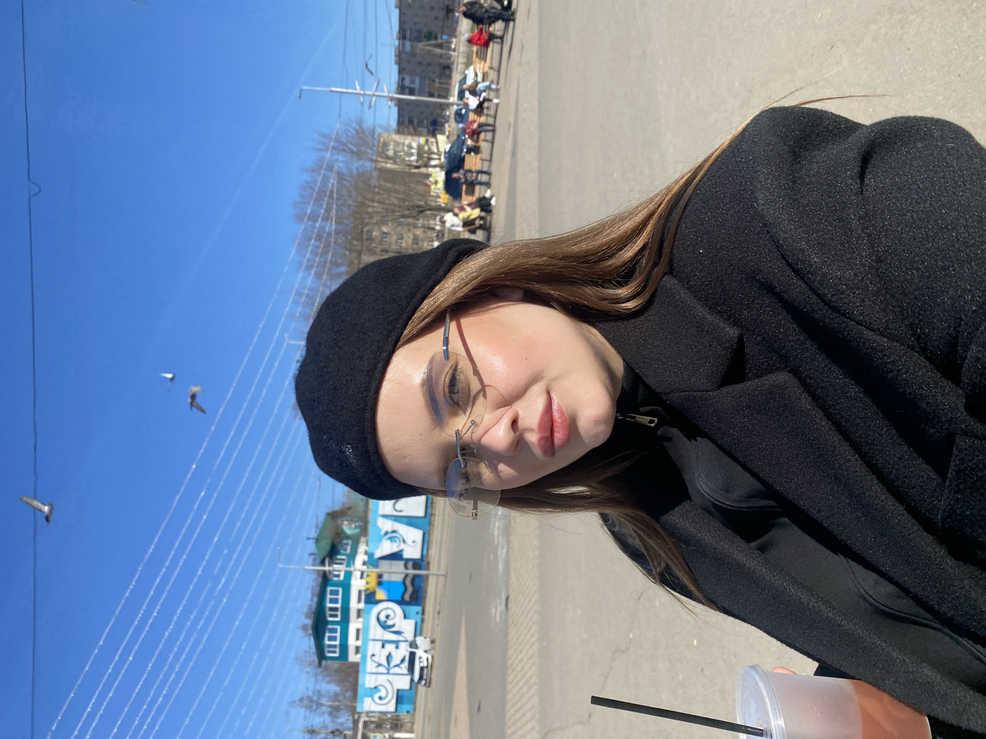 A young woman is depicted in a close-up, outdoors on a bright, sunny day. She has long brown hair, wears a black beret, a black coat, and rimless glasses. She holds a clear plastic cup with a dark straw, containing a light-colored beverage. Her face is turned slightly towards the camera, with a calm, almost smiling expression.
The background features a clear, vivid blue sky with multiple thin lines stretching across it, likely utility wires. Three birds are visible flying in the distance. To the left of the woman, a distinctive building with a blue and white painted facade stands out. The facade displays large, stylized letters that appear to spell "ЧЕРН", consistent with the provided location of Chernushka, Russia. In the further distance, other buildings and bare trees are visible, suggesting a non-summer season.
To the right, a paved ground area extends back, where several people can be seen seated on benches, indicating a public pedestrian space. The strong sunlight creates clear shadows, suggesting it is midday. The scene captures a casual outdoor moment in an urban setting.