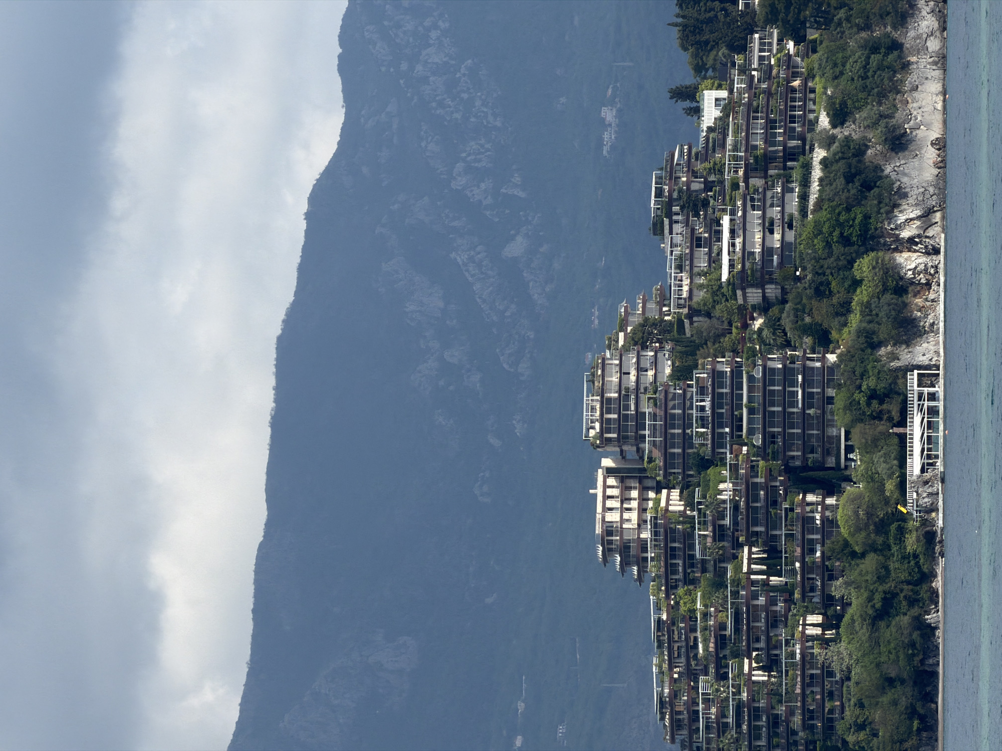 This image captures a serene coastal landscape featuring a modern architectural complex nestled into a hillside. The scene is dominated by a large body of calm, turquoise water in the foreground, bordering a rugged, rocky coastline.
Perched on the steep green slope above the water is a sprawling complex of terraced, multi-story buildings. These structures exhibit contemporary architecture with extensive glass facades, large balconies, and a blend of white and darker materials. Lush green vegetation, including trees, bushes, and plants integrated into the balconies, heavily adorns the hillside and the buildings, creating a harmonious blend with the natural environment. The buildings appear to cascade down the slope towards the sea, offering panoramic views.
In the background, a vast, dark mountain range rises dramatically, displaying rocky textures and sparse vegetation under an overcast sky filled with soft, diffused clouds. The overall lighting suggests it is daytime, but without direct sunlight.
No people or specific activities are visible, lending a tranquil and pristine atmosphere to the scene. There is no visible text in the image. Given the location context, this scene is characteristic of luxury coastal developments along the Budva Riviera in Montenegro, such as Dukley Gardens.