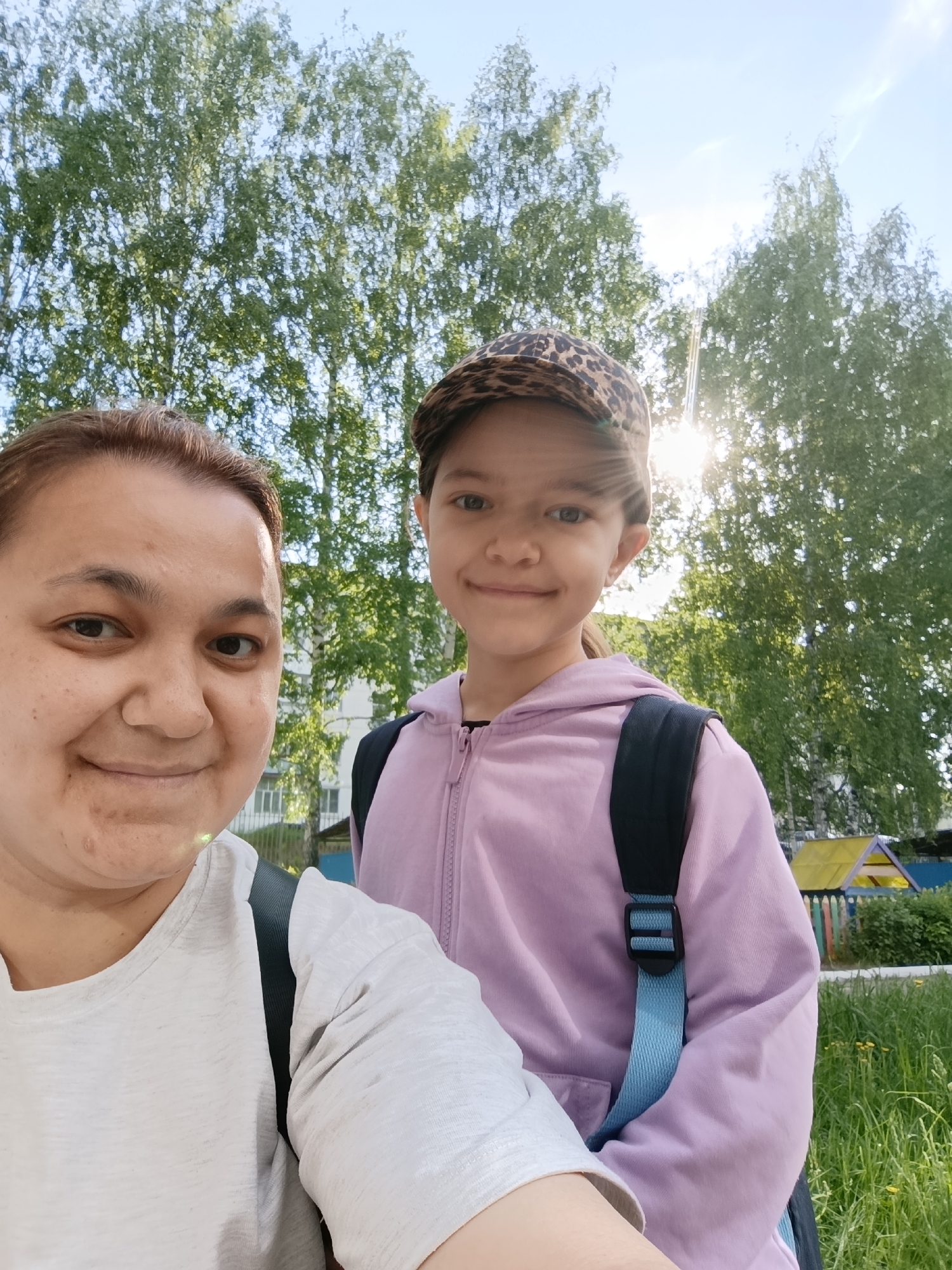 A woman and a young girl are posing for a selfie outdoors. The woman is on the left side of the frame, with fair skin and short brown hair pulled back. She is wearing a light-colored t-shirt with a black strap from a backpack visible on her shoulder. The girl is to the right of the woman. She has light skin, a slight smile, and a cheetah-print baseball cap. She is wearing a pink hoodie and a black backpack with a light blue strap.
The background includes trees with lush green foliage, the sun shining through the trees. A building is partially visible behind the pair. The image appears to be taken in an urban area, possibly a park or residential neighborhood.
Given the information in the prompt, it can be inferred that the location is in the city of Chaikovsky, Russia.