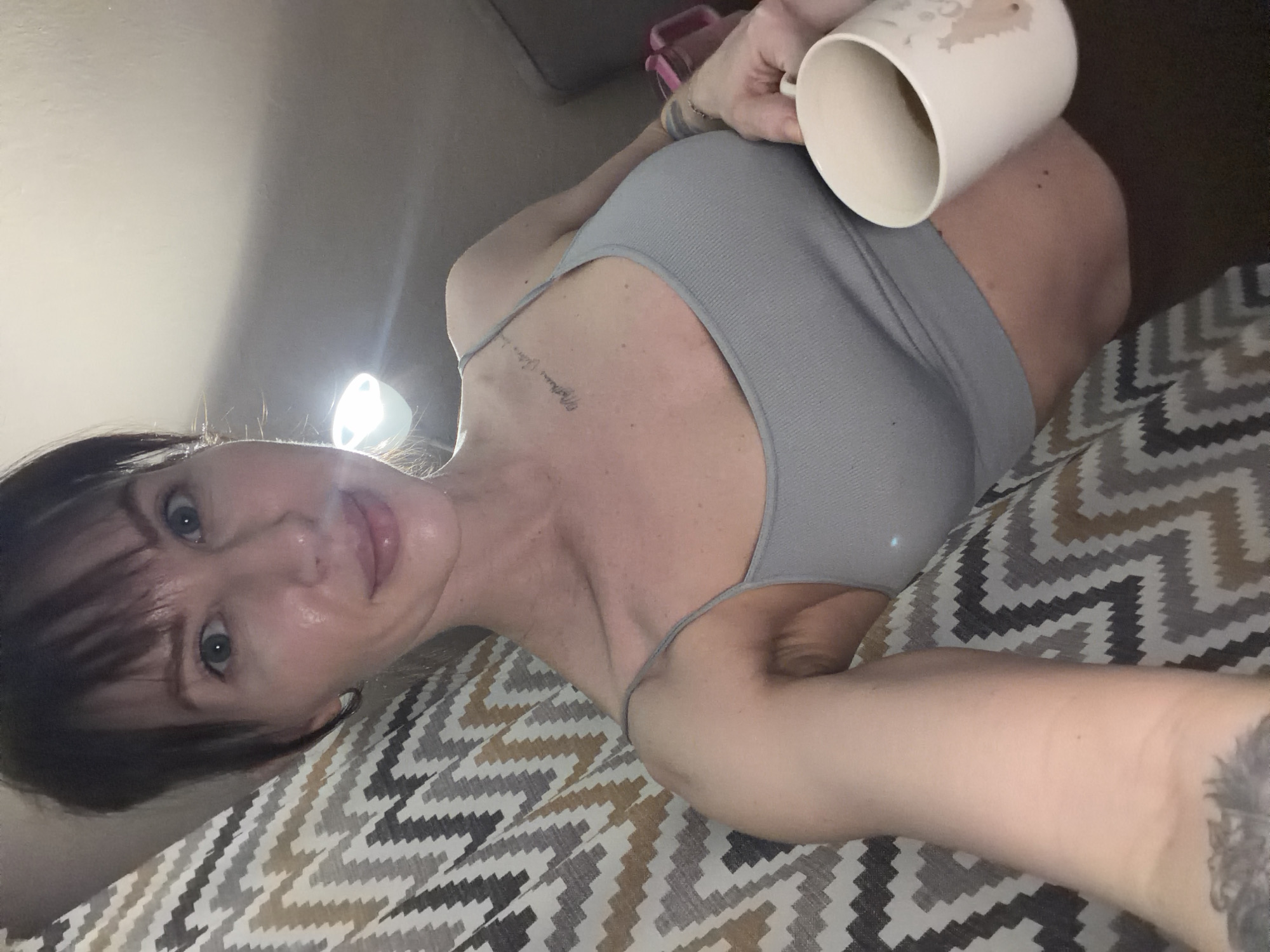 A woman with dark hair and bangs is lying on a chevron-patterned bedspread, holding a white mug in her right hand. She is wearing a gray tank top. There is a light source shining behind her head, causing a glare. A glimpse of a pink object is visible in the background. Tattoos are visible on her left arm and near her collarbone. There is no indication of the location being Perm, Russia, in the image.