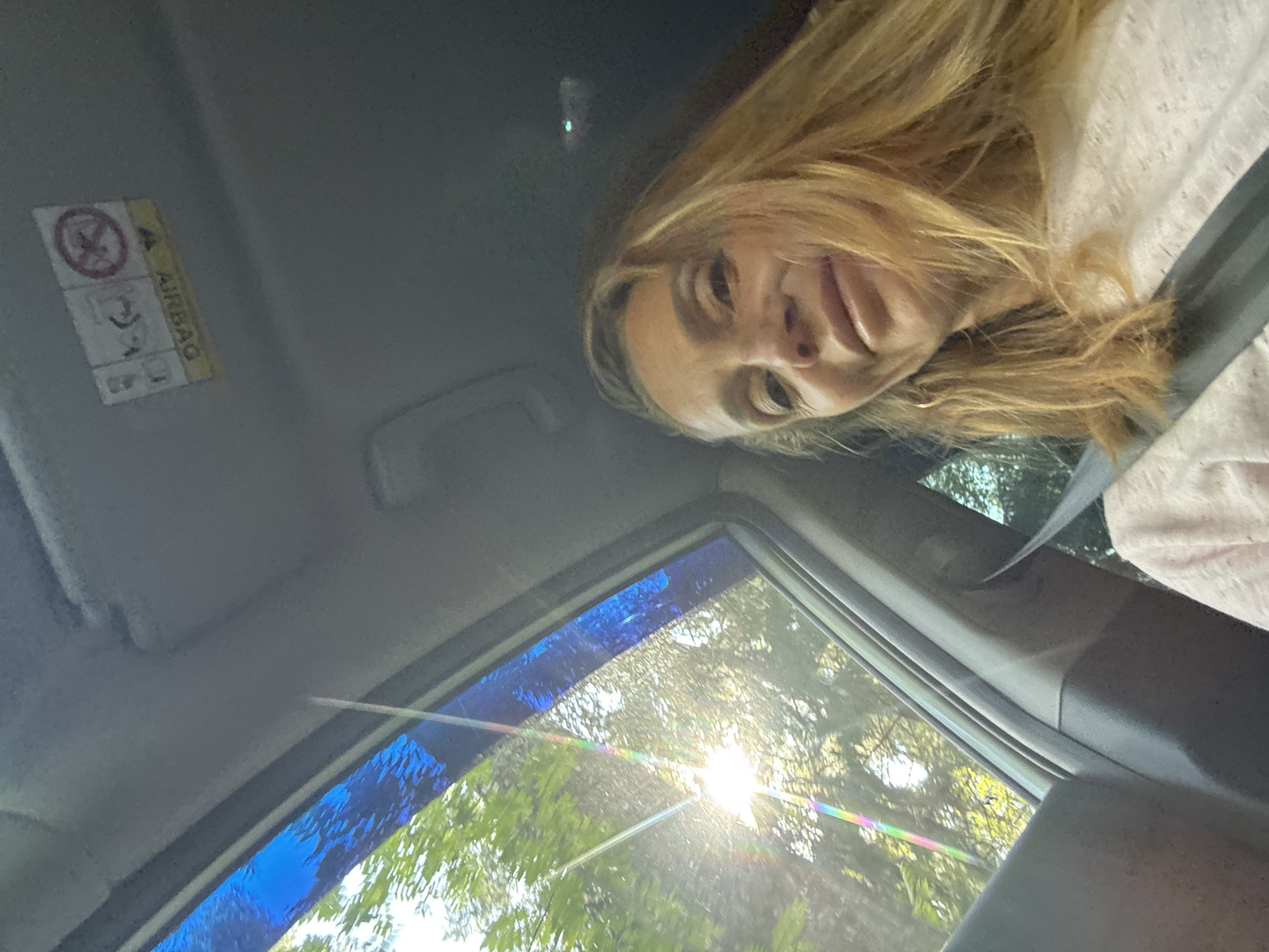 A woman with blonde hair and a pink shirt is in the passenger seat of a car. She is looking at the camera. The sun is shining through the window, casting a rainbow effect on the car's interior. The car is in the city of Portovoe, Russia.