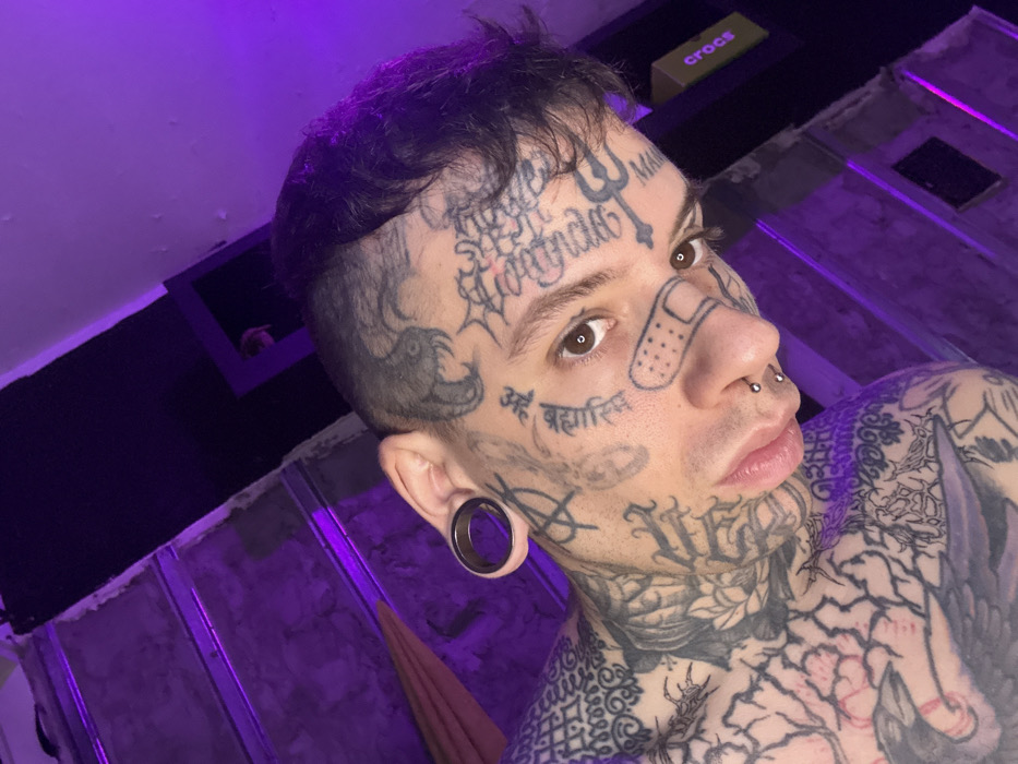 This close-up, angled portrait depicts a young adult male in an indoor setting within Buenos Aires, Argentina. The subject, positioned with his head slightly tilted and looking directly at the camera, exhibits extensive black ink tattoos covering his face, neck, and visible chest/shoulder areas. Notable facial tattoos include a stylized adhesive bandage on his left cheek, Devanagari script on his left cheek, an 'A' with a circle over it near his left ear, a spiderweb on his right temple, a snake on his upper right forehead, and a trident-like symbol on his right forehead. He wears a black ear gauge in his left earlobe, a bridge piercing, and a double nostril piercing with silver studs.
The background is illuminated by a dominant purple ambient light. To the left, a section of a light-colored staircase or stepped structure is visible, alongside a draped brownish fabric. To the right, a plain wall displays a dark rectangular frame, with a yellow "Crocs" box featuring black text positioned on a surface above it. The scene suggests an interior space with a functional or unadorned aesthetic, characterized by the artificial purple illumination.