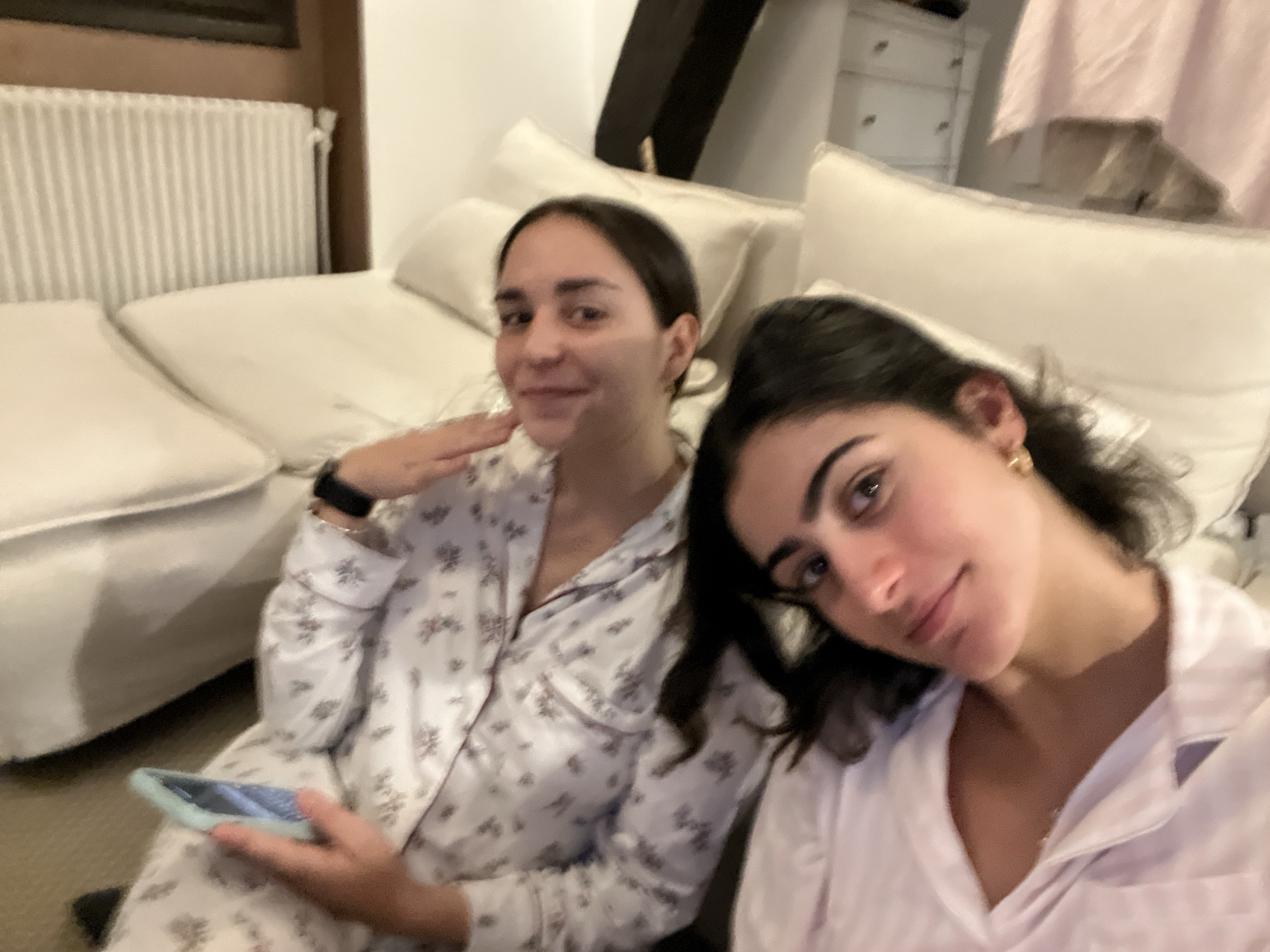 Two young women are sitting on a white sofa, wearing pajamas. The woman on the left holds a phone and has a watch on her wrist. The background includes a radiator, a shelf, and a cabinet. The location is not discernible as Paris, France, based on the provided image.