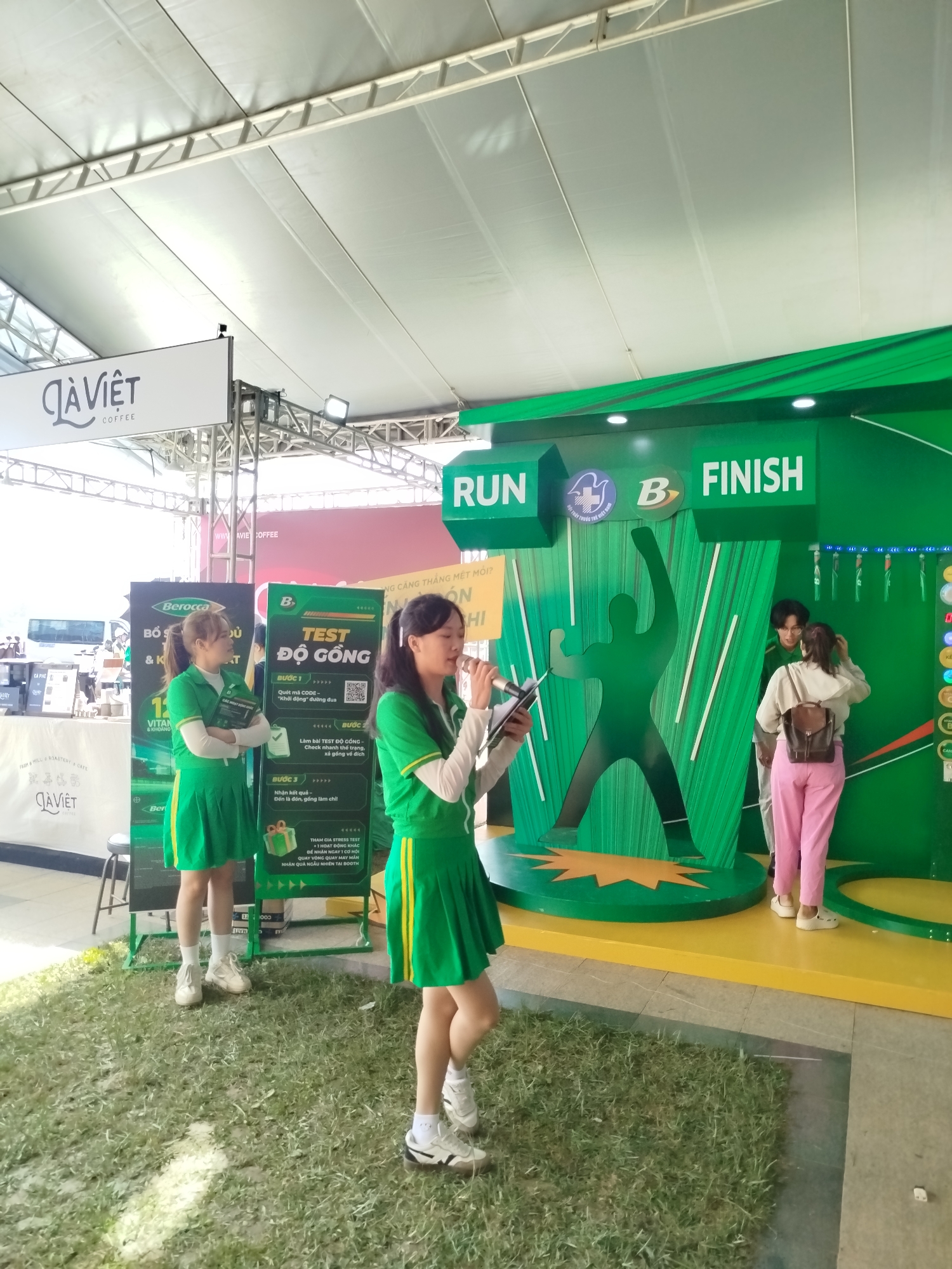 This image depicts a lively promotional event taking place under a large white tent structure, likely in an outdoor or semi-outdoor setting in Da Lat, Vietnam, during the daytime.
The primary focus is a vibrant green-themed booth prominently featuring "RUN B FINISH" and the "Berocca" brand logo, indicating a health or wellness product promotion. Two female staff members, dressed in matching green polo shirts with yellow stripes and pleated green skirts, are central to the activity. One staff member on the left holds a product box, while the other in the foreground holds a microphone and a small device, actively engaging with the audience or facilitating an activity.
The main green booth advertises a "TEST ĐỘ GỒNG" (Resilience Test or Stamina Test). Signs detail the participation steps: "BƯỚC 1: Quét mã CODE - "Khởi động" đường đua" (Scan code to start the race), "BƯỚC 2: Làm bài TEST ĐỘ GỒNG - Check nhanh thể trạng, xá gồng về đích" (Perform the test to quickly check physical condition and push to the finish), and "BƯỚC 3: Nhận kết quả" (Get results). Participants are informed that by joining the "STRESS TEST + 1 HOẠT ĐỘNG KHÁC," they get a chance to spin a lucky wheel and receive a random gift. A large green silhouette of a person with arms raised, symbolizing strength or victory, is a key visual element of the booth.
Several attendees are present, including a couple interacting near the "FINISH" section of the green booth. The ground in the foreground is covered with artificial grass, enhancing the outdoor event feel. To the left, another booth for "QÀ VIỆT COFFEE" (Vietnamese Coffee) is visible, suggesting multiple vendors or activities at the event. The overall atmosphere appears engaging and energetic, focused on promoting health and physical well-being.