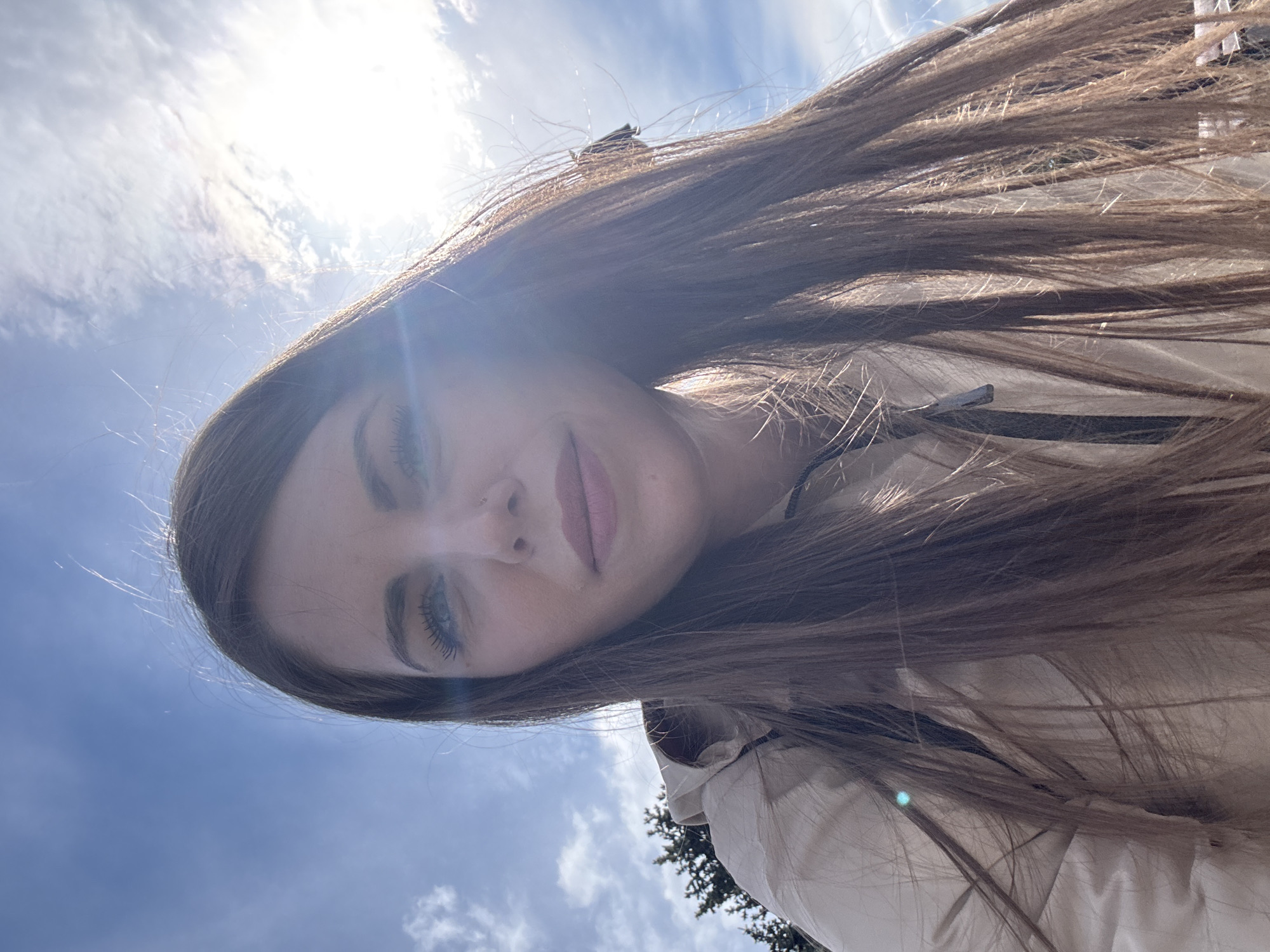 A woman with long brown hair is centered in the image. The background shows a blue sky with some clouds. The sun's glare obscures some details. A beige-colored jacket is visible on the right side of the image. There is a tree in the lower right corner. No other details regarding Moscow, Russia, or specific locations, subjects, objects, actions, or interactions are apparent.