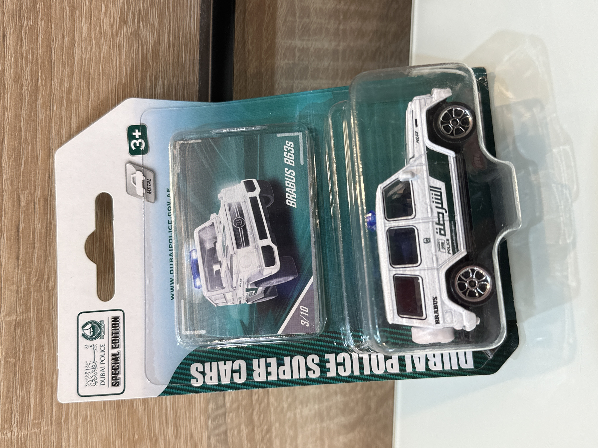 The image shows a toy car, a Brabus B63S Dubai Police special edition, still in its packaging. The packaging indicates it's a metal toy suitable for ages 3 and up. The car itself is white with green accents and the word "POLICE" and "الشرطة" (Arabic for "The Police") written on it. The packaging also shows the Dubai Police logo and website address, as well as the edition number, 3/10. The toy is resting on a surface, possibly a table, with a wooden background to the left and a white surface to the right. There is nothing indicating the city of Prisovo, Bulgaria.