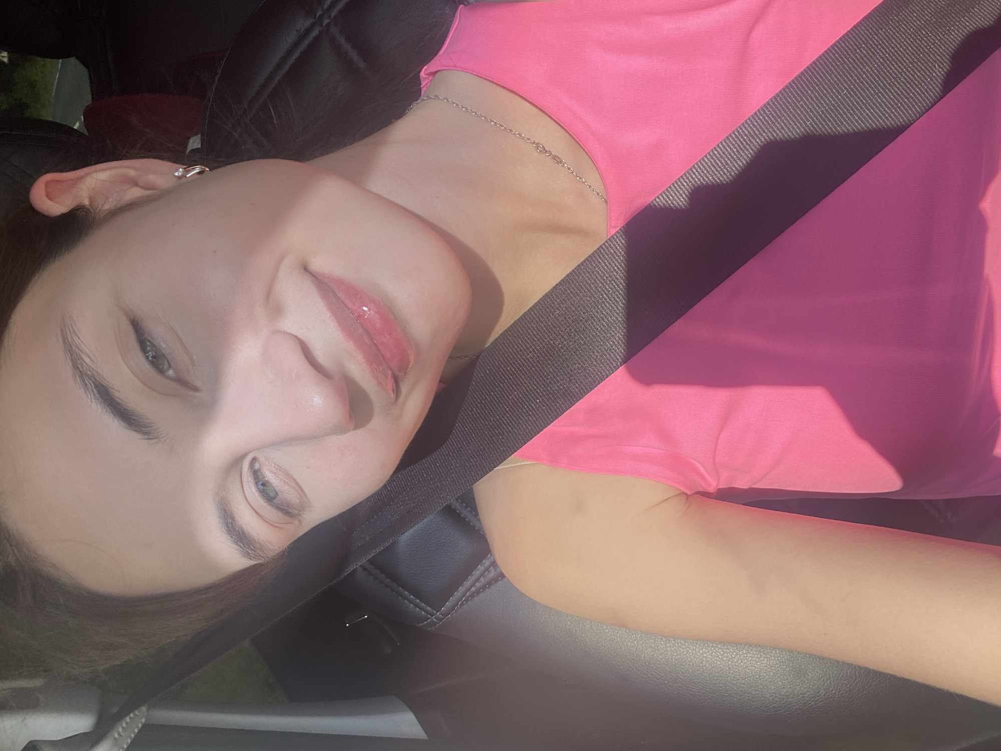 A young woman with light skin, dark brown hair, and green eyes is pictured in a car. She is wearing a pink tank top and a silver necklace. Her eyes are looking towards the viewer with a slight smile on her face, and she has lip gloss on. A seatbelt is visible across her chest. The background of the image is blurred, showing the interior of a car with leather seats and a road visible through the window. The sun is casting shadows on her face and clothing. The location is identified as being in the city of Kamkoye, Russia.