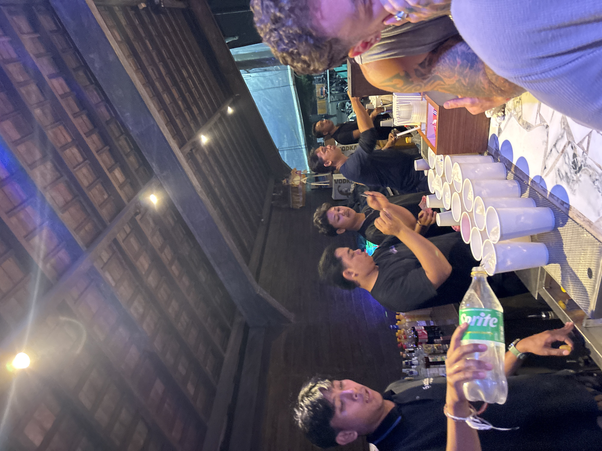 In North Kuta, Indonesia, four men are visible behind a bar. The man on the far left, wearing a black polo shirt, is pouring Sprite from a plastic bottle into a white paper cup. In the center, another man in a black t-shirt is holding a white paper cup and appears to be gesturing with his other hand. To his right, a third man in a black t-shirt is also behind the bar. On the far right, a partially visible man with tattooed arms is in the foreground, looking towards the bar. The bar is stocked with various bottles of alcohol, and there are rows of white paper cups on the counter. The ceiling above the bar is constructed with wooden beams and tiles, illuminated by bright lights.