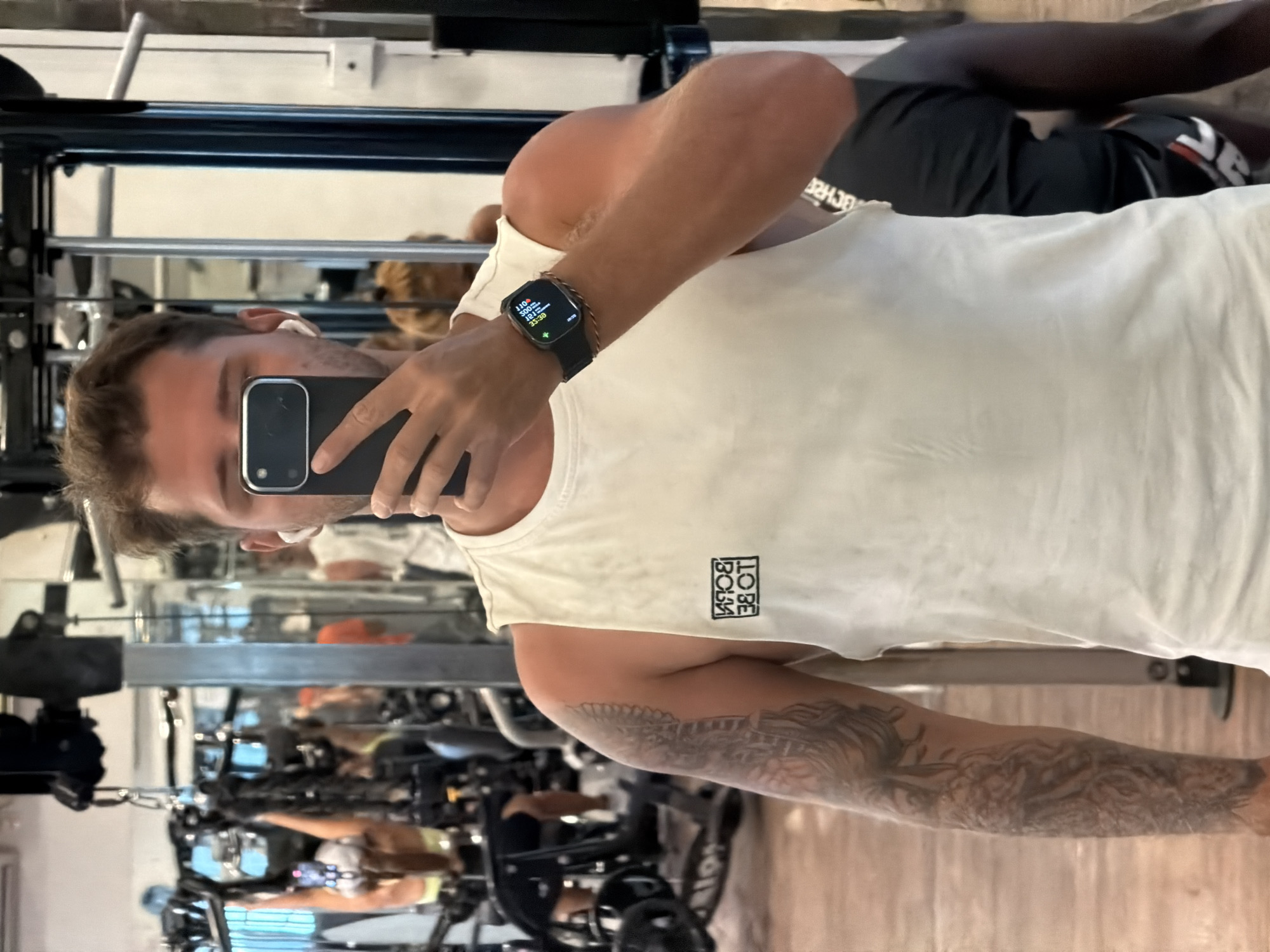 This mirror selfie, presented rotated 90 degrees clockwise, features a man in a gym setting in Denpasar Barat, Indonesia. The main subject, visible from the chest up, has light brown hair and a prominent sleeve tattoo on his left arm. He is wearing a white tank top with a black square logo on the left chest that, when unmirrored, reads "TO BE BORN". He holds a black smartphone with multiple lenses, obscuring most of his face, to capture the image. On his right wrist, a black smartwatch displays fitness metrics including a heart rate of "110BPM", "182KCAL" burned, and a workout duration of "35:36", alongside the time "05:36".
The reflection in the large mirror reveals the interior of a gym with various exercise machines and a wooden floor. Several other individuals are visible in the background reflections, seemingly engaged in workouts. One person, partially obscured and appearing to be lying or bent over, wears black athletic clothing. The overall scene depicts a typical indoor fitness environment.