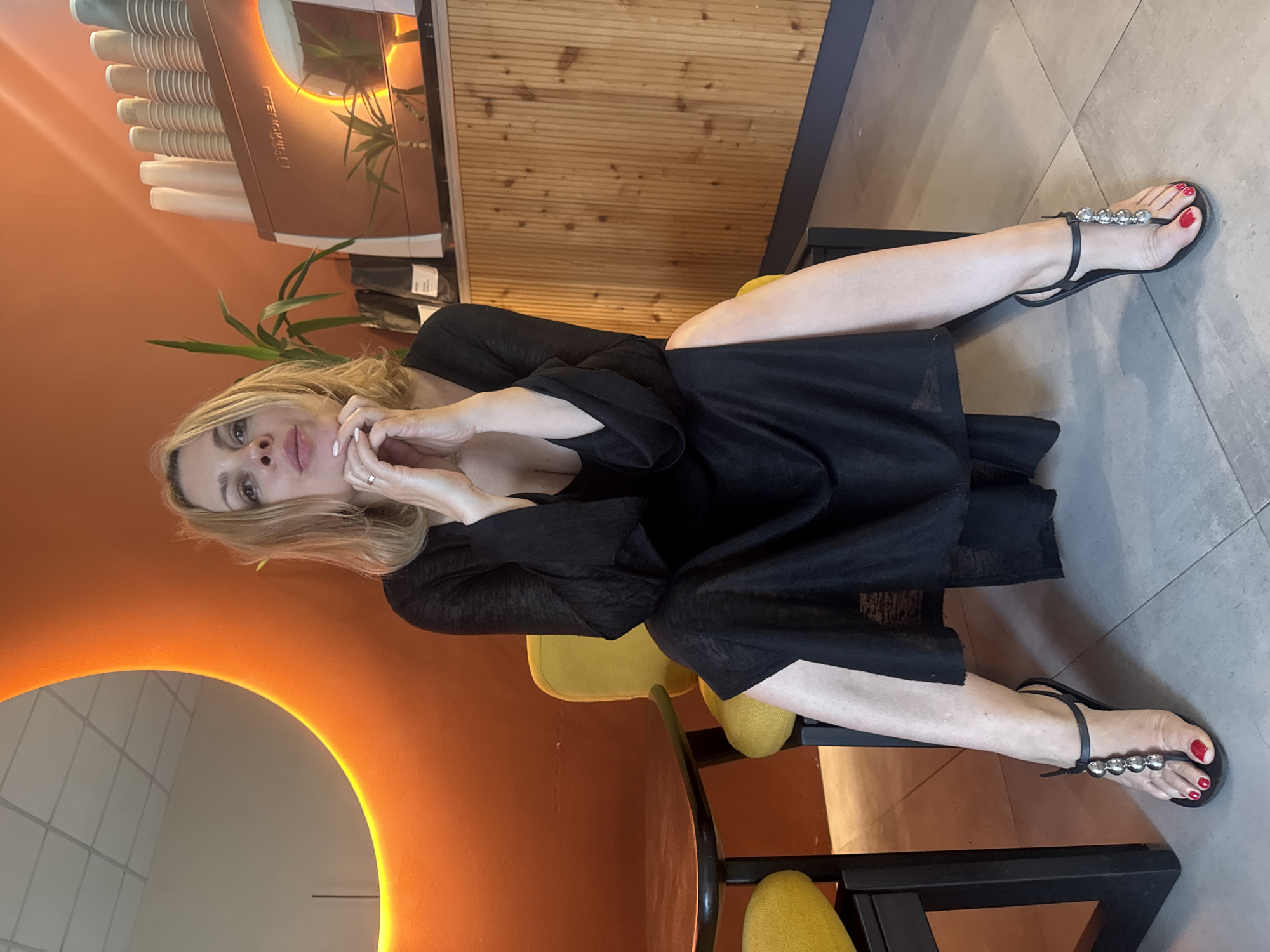 A woman with blonde hair sits on a yellow chair in what appears to be a cafe. She is wearing a long, black, flowy dress and black sandals with silver embellishments. Her hands are clasped in front of her chin, and she is looking towards the camera. In the background, there's a large orange wall with a crescent-shaped mirror illuminated with a yellow light, a coffee machine, cups, and plants. The floor is tiled. The scene is indoors, and the lighting suggests it is daytime. The woman's legs are crossed, and her feet are on the floor.
