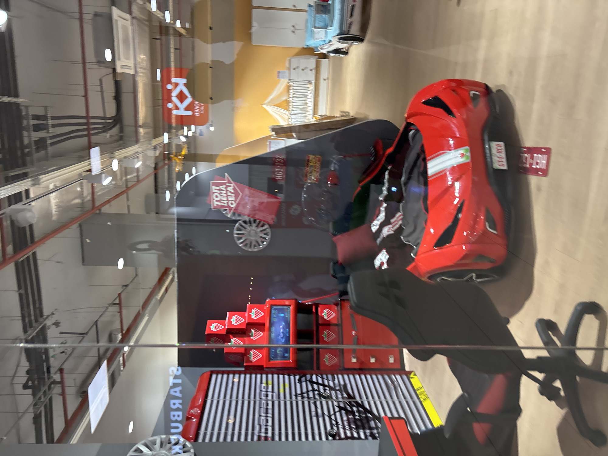 The image shows a display with various items, including a red car model, strawberry-themed boxes, and a digital screen, possibly located inside a retail establishment in Plovdiv, Bulgaria. This is determined from the Cyrillic text "STARBUCKS" visible on the display and the license plate "HGT-132" on the car model. The environment is an indoor space with industrial-style ceilings and visible structural support. There are no people directly visible in the image.