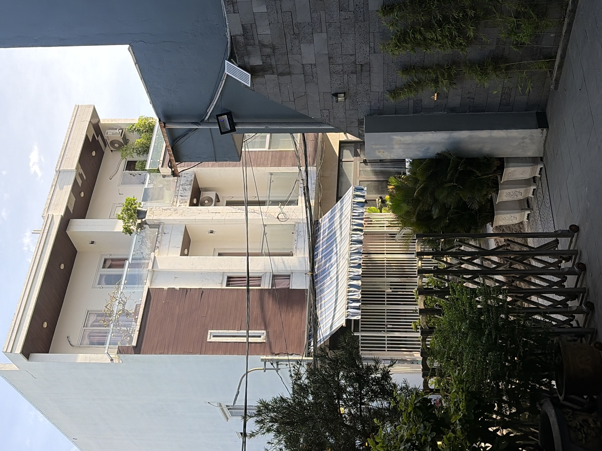 This image depicts a street view in Da Nang, Vietnam, featuring a multi-story building with a white facade and brown wooden accents. The building has multiple balconies with glass railings, some adorned with potted plants. Visible air conditioning units are mounted on the exterior walls. In the foreground, a metal accordion gate encloses an area with potted plants and a small tree. Adjacent to the building, a grey wall constructed of large stone blocks features a solar panel and outdoor lighting fixtures. The sky above is partly cloudy.