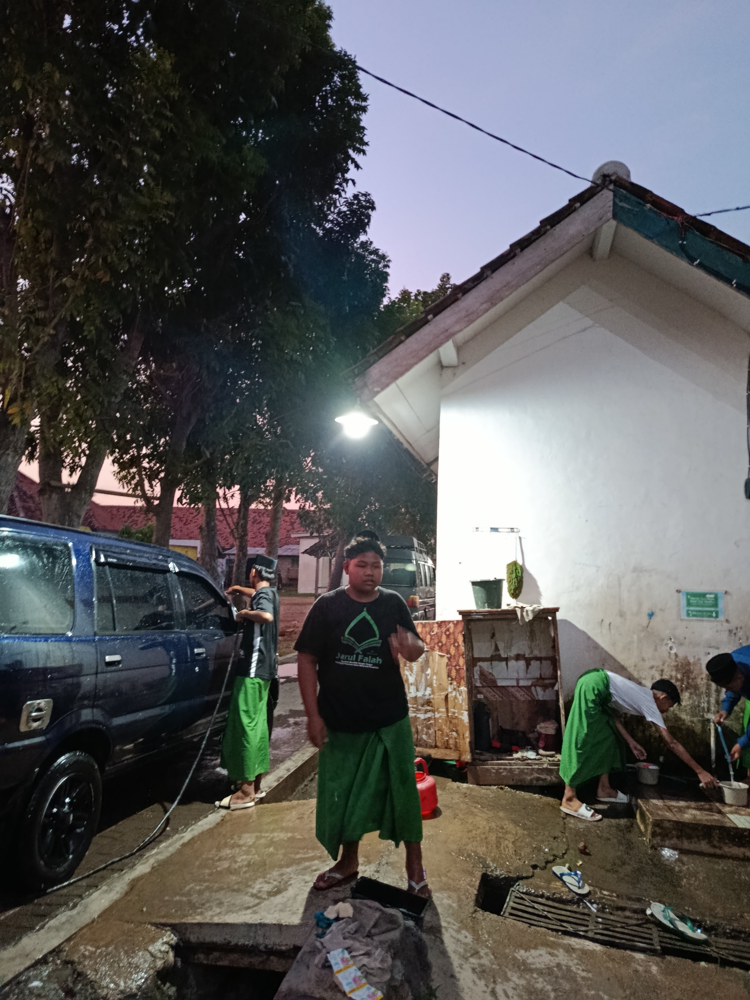 At dusk in Ramban Wetan, Indonesia, a group of young men are gathered around a car wash area outside a building. A dark blue SUV is parked to the left of the frame, and a man in a black t-shirt with green trim and a black skullcap is washing it with a hose. He is wearing a green sarong. In the foreground, a young man in a black t-shirt that reads "Jarun Falah" and a green sarong is gesturing with his right hand. To the right, two other young men, also wearing green sarongs, are washing their hands and faces at a concrete sink. The building behind them has a white facade with a sloped, tiled roof and an outdoor sink area. Trees with dense foliage are visible in the background, and the sky is a soft twilight blue.