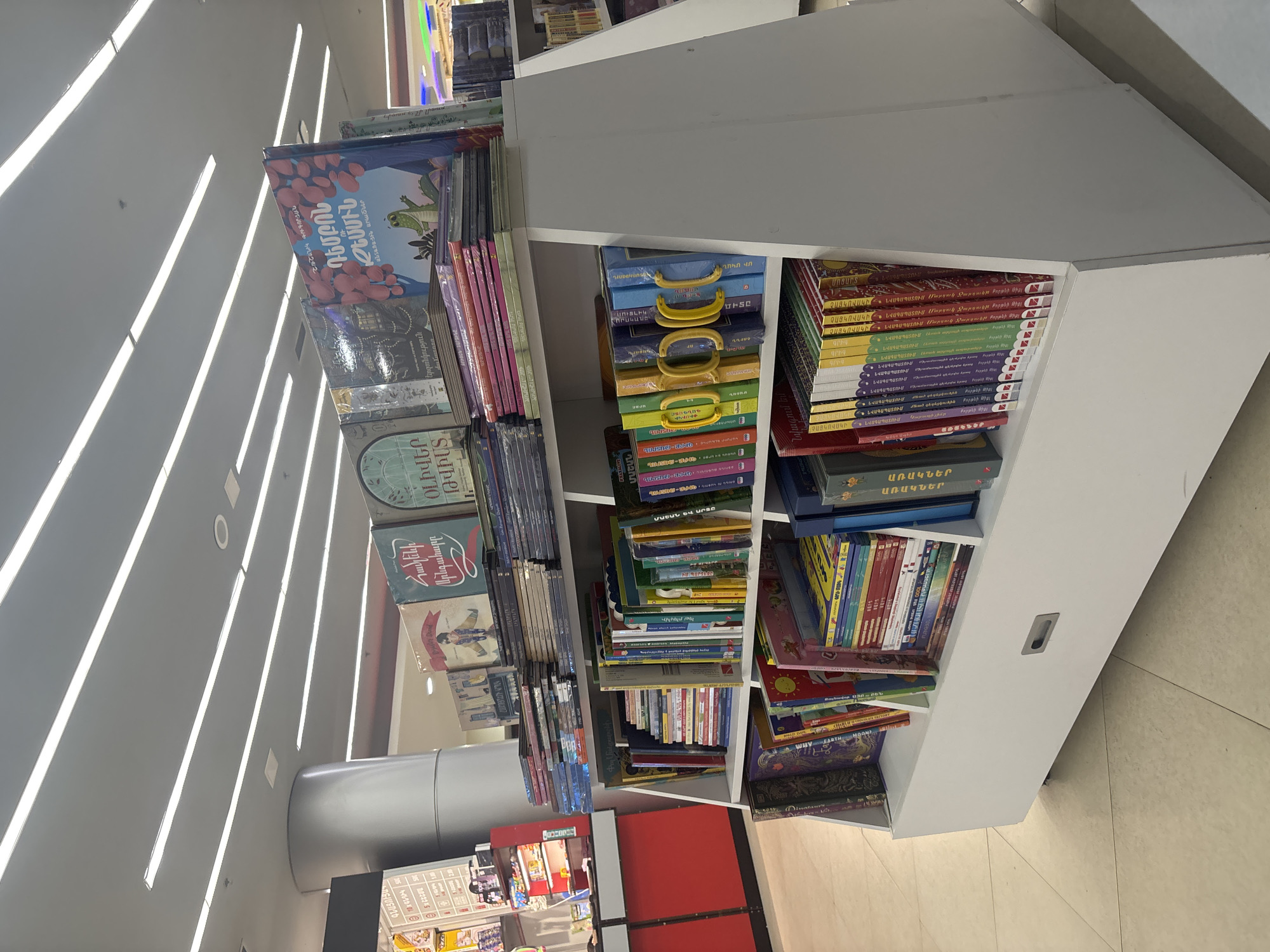 The image displays an interior view of a modern bookstore in Yerevan, Armenia, characterized by bright, even lighting and white shelving units. The primary subjects are numerous books, organized neatly on these shelves.
Books are presented in various configurations: many are stacked spine-out, others are face-out to showcase their colorful covers, and some children's book sets are packaged in box-like containers with handles. The overall aesthetic is clean, organized, and inviting. The floor features light-colored tiles, reflecting the overhead fluorescent lighting strips.
Visible text on the book covers and spines is predominantly in Armenian, supporting the given location context. Identified titles and words from the OCR include "VANSK," "ԳՆԱՑՈՒՑԱ" (Price List, visible on a children's book with an alligator), "Արնգահարը ՕԼԻՎԵՐ" (likely related to Oliver), "Համշենի" (of Hamshen), "ԴԵՄԲՈՆ" (Dembon), "XATALOVE," "ՄԱՇԱՆ" (Mashan), "երջանկություն" (happiness), "ՄԻ ՄԿԱՆ" (One Mouse), "ԳԱՆԳԱՏԸ" (The Complaint), and "Նվագում եմ" (I play/I'm playing).
A significant portion of the collection appears to be children's books or adaptations, with multiple volumes labeled "ՆՎԱԳԱՊԱՏՈՒՄ" (Play/Performance or Adaptation), often associated with "ԿԱՊՈՒՏԱԿԱՆ" (Kaputakan), possibly a series or publisher. Specific examples include "ՉԱՅԿՈՎՍԿԻ ՆՎԱԳԱՊԱՏՈՒՄ Կարապի լիճը" (Tchaikovsky's Swan Lake Play/Performance) and "ՀՈՒՄՈՐՍՏԻՆ ՆՎԱԳԱՊԱՏՈՒՄ" (Humor's Play/Performance). Other books are identified as "ԱՌԱԿՆԵՐ" (Fables).
No people are visible in the frame, suggesting a quiet moment within the store. The time of day is not ascertainable due to the reliance on artificial lighting. The scene conveys an atmosphere of order and accessibility for readers.