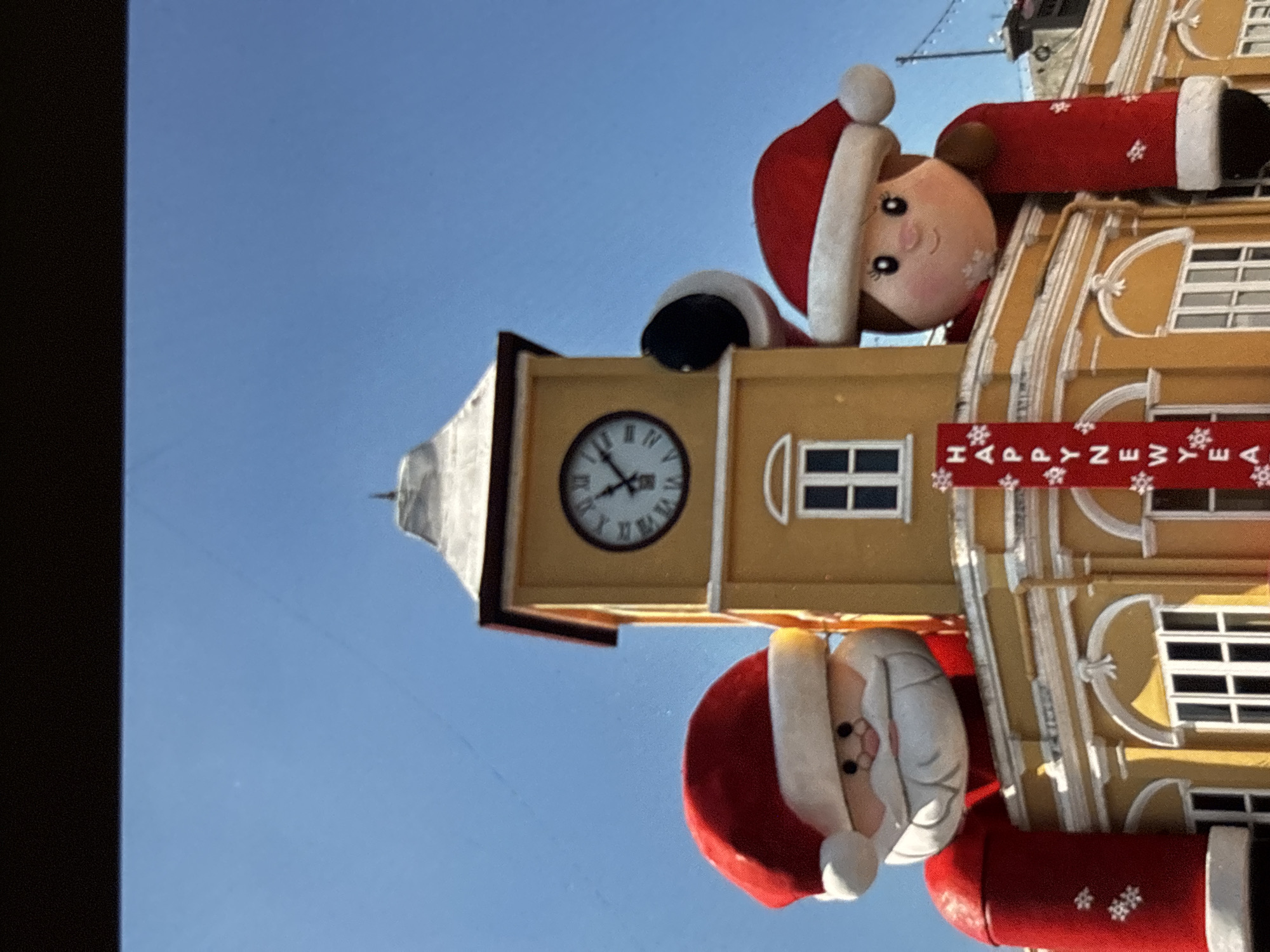 A photograph captures a bright, sunny day with a clear blue sky, featuring a yellow clock tower building. Two large, inflatable figures, one resembling Santa Claus and the other a female character in festive attire, are positioned on top of the building, peeking over its edge. The Santa figure has a white beard and a red hat, while the female figure also wears a red hat and a red garment with white trim. A banner with the words "HAPPY NEW YEAR" and snowflake decorations hangs vertically on the building. The building itself is adorned with white architectural details and multiple windows. The scene suggests a celebratory atmosphere, likely during the holiday season.
