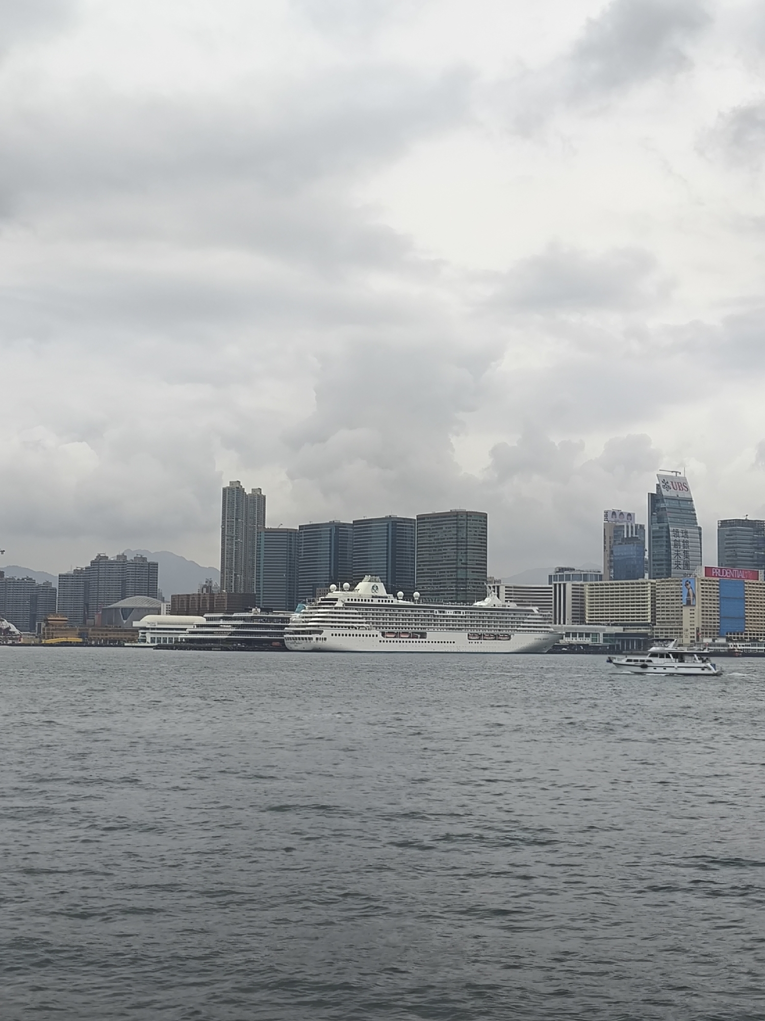The image depicts Victoria Harbour in Hong Kong, China, under an overcast sky filled with dense, uniform grey clouds. The foreground is composed of dark, choppy water with visible ripples. In the mid-ground, a large white cruise ship, identified as "CRYSTAL SYMPHONY" by lettering on its bow, is docked at a pier. It features multiple decks and a green logo on its funnel. To the right of the cruise ship, a smaller white motor yacht is observed in motion on the water, moving leftwards and creating a subtle wake. The background displays a dense urban skyline consisting of numerous high-rise buildings, including commercial towers with glass facades and residential structures. Identifiable signage on buildings includes "UBS" on a prominent skyscraper and "PRUDENTIAL" on another building. A distinctive domed structure is visible behind the cruise ship. Distant mountains are faintly discernible behind the city's architecture. No individuals are clearly visible.