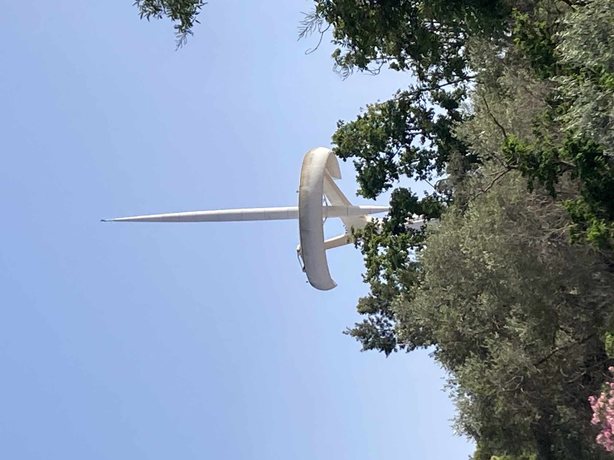 This image shows the Montjuïc Communications Tower, also known as Torre Calatrava, in Barcelona, Spain. The tower, designed by Santiago Calatrava, is a prominent landmark on Montjuïc hill, visible against a clear blue sky. The tower is characterized by its sleek, white, needle-like structure topped with a spire. In the foreground, lush green trees and foliage partially obscure the base and middle sections of the tower, suggesting it is surrounded by parkland or a natural setting. The trees are diverse in type and density, with some bearing dark green leaves and others a lighter, silvery-green hue. The overall impression is that of a sunny day, with the tower standing tall amidst a verdant landscape in the city.
