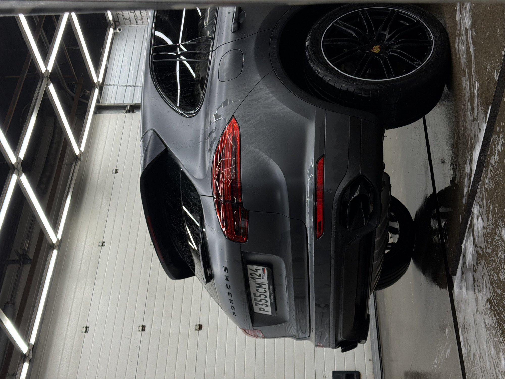 The image shows the rear passenger side of a dark gray Porsche Cayenne parked in a dimly lit garage. The vehicle is wet, indicating it may have recently been washed or is being detailed, with water droplets visible on its surface. The license plate, which appears to be a Russian plate from Krasnoyarsk (indicated by "RUS" and the format of the alphanumeric characters "P355CM 124"), is clearly visible. The garage has a white, corrugated metal door in the background and overhead fluorescent lighting. The floor is wet and reflects the car and lights, with some foam or soap suds visible. The overall environment suggests a car wash or detailing bay.