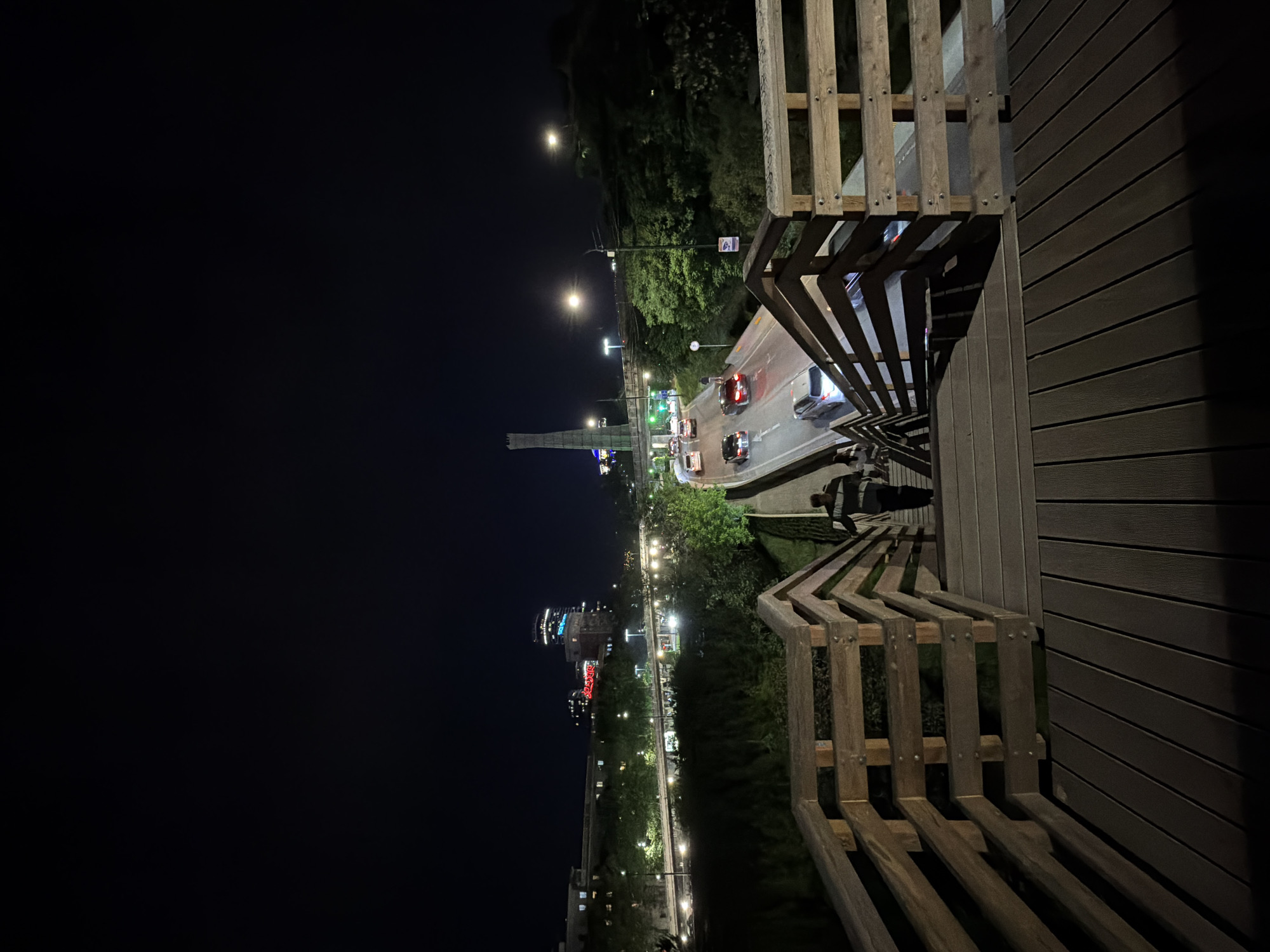 The image depicts a nighttime view overlooking a street in Tbilisi, Georgia, from an elevated wooden walkway with railings. Cars are visible on the street, their headlights and taillights creating streaks of light. A tall, illuminated structure is visible in the distance, and buildings with lights on are seen along the street. Trees line the street. The night sky is dark. A person can be partially seen on the walkway. The immediate foreground consists of the wooden walkway and railings.