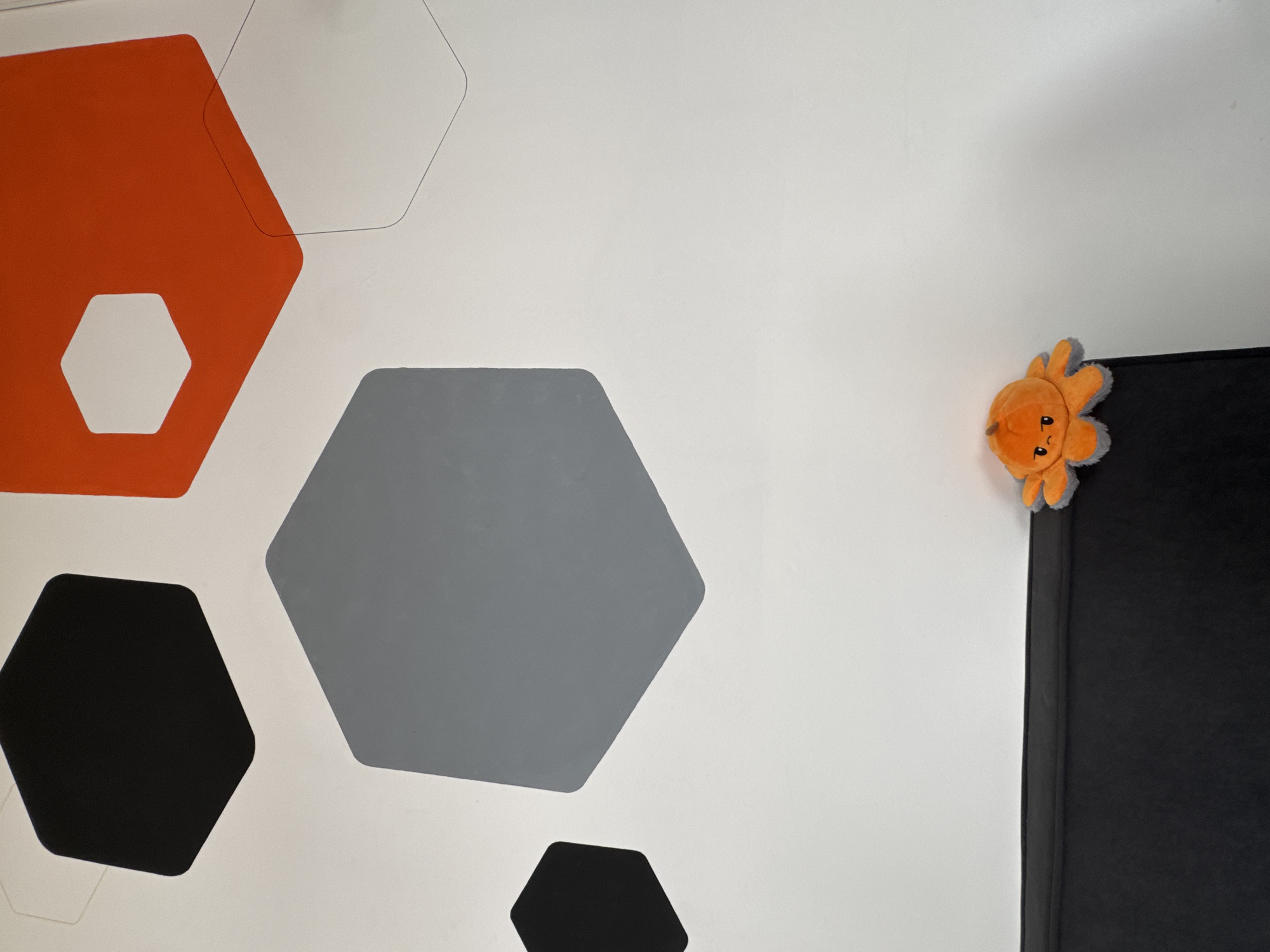 This is a close-up, eye-level shot of a wall with a hexagonal geometric pattern and a plush toy. The wall is painted white and features several large hexagons in black, gray, and orange. Some hexagons have a light gray outline, indicating they are part of the pattern but not filled in. In the lower right quadrant of the image, on top of a black surface, sits a small, orange plush toy resembling an octopus. The toy has a frowning face with gray trim around its tentacles. The scene appears to be indoors, likely in a room. There is no indication of the location being in Kaliningrad, Russia.
