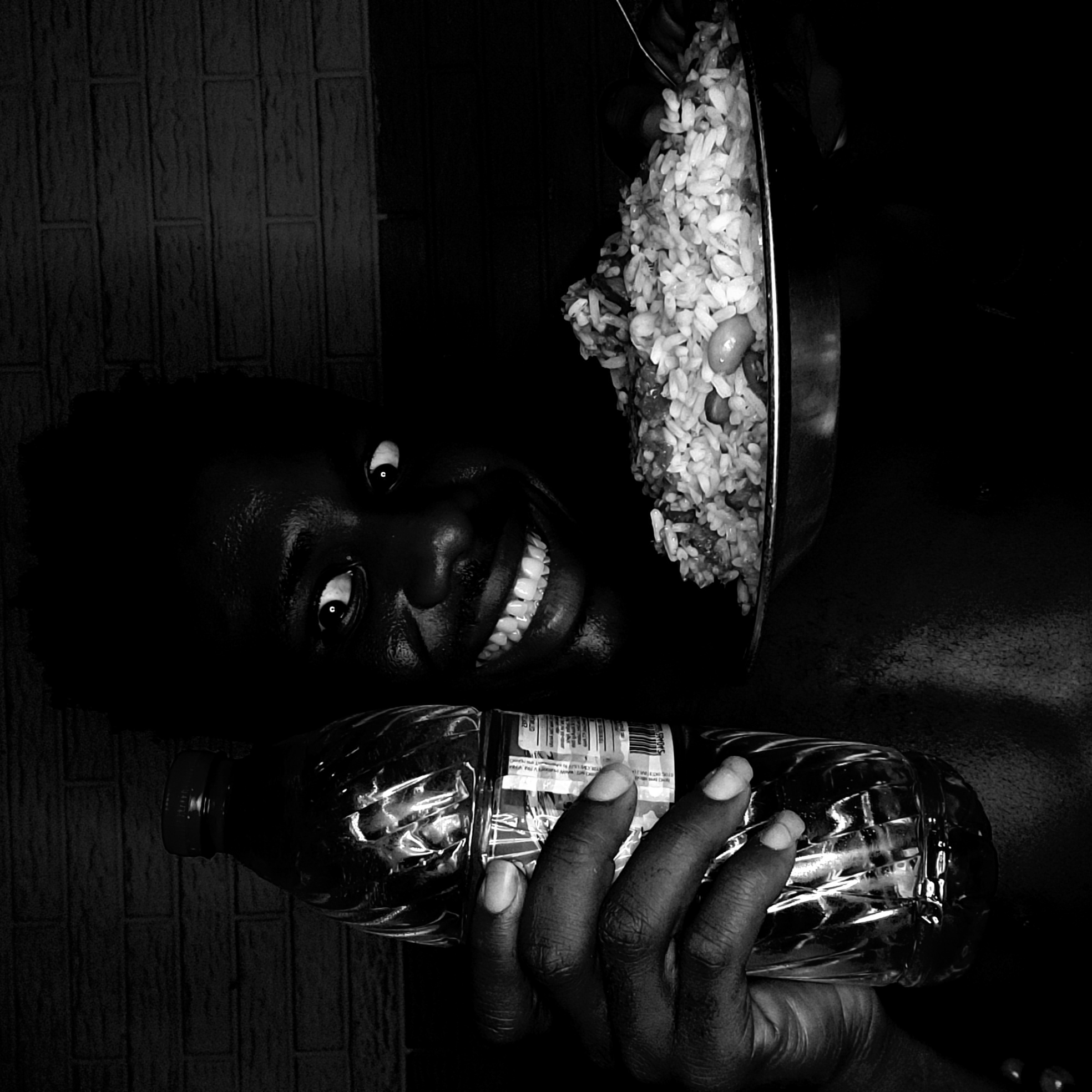 A black and white photograph, likely taken indoors in Narayi, Nigeria. A person with dark skin is holding a clear plastic water bottle in their hand. Next to their head, a pan filled with rice and beans is visible. The background features a textured wall. The person is smiling, looking directly at the camera.