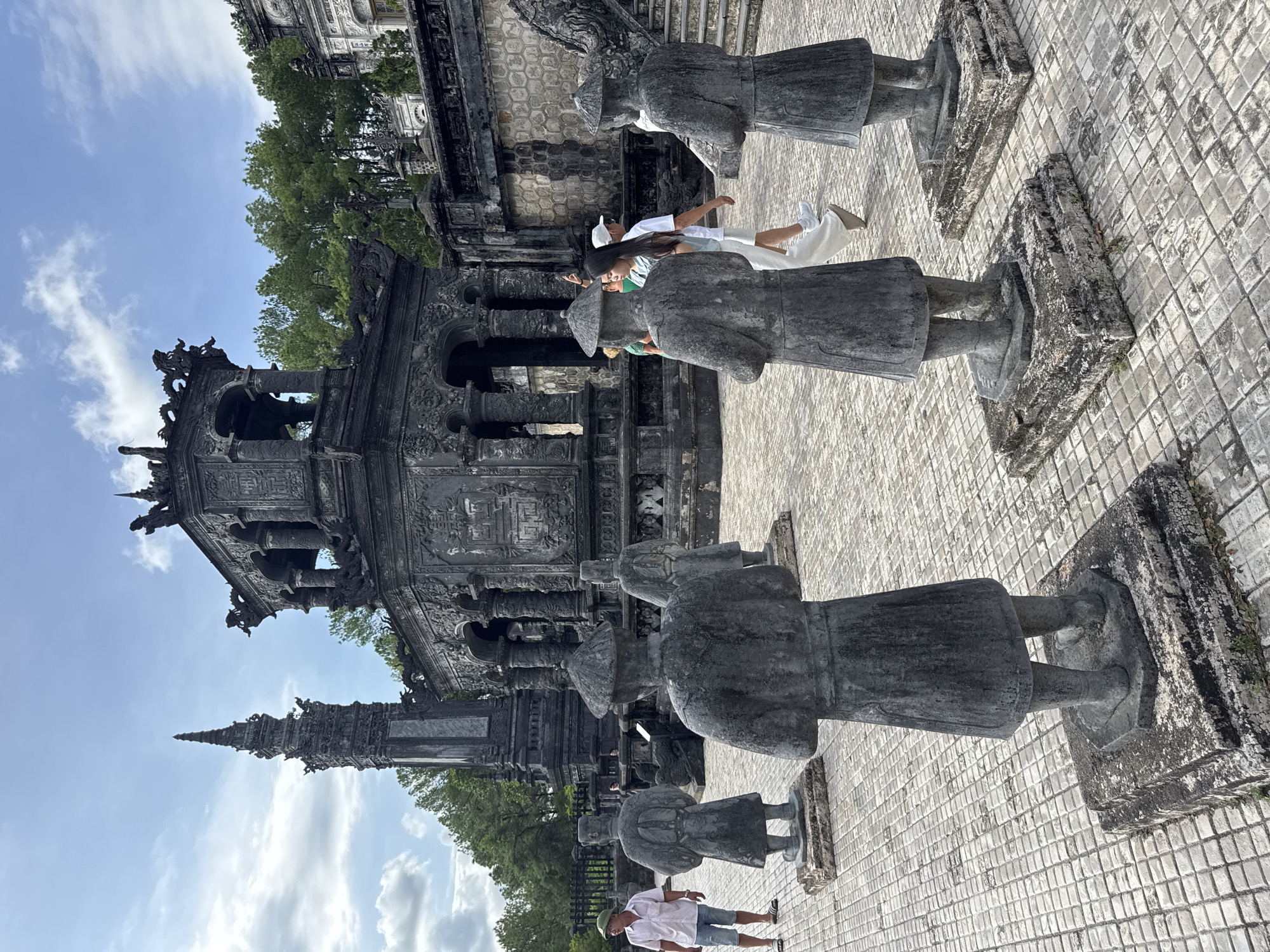The image shows the exterior of Khai Dinh Tomb, located in Thủy Xuân Ward, Vietnam. The tomb is characterized by its dark, ornate architecture, featuring intricate carvings and dragon motifs. In the foreground, four stone statues of mandarins stand in a line, facing away from the viewer. These statues are weathered and appear to be made of granite. Behind them, the main structure of the tomb is visible, with its tiered roofs and elaborate decorative elements. A tall, slender tower stands to the left of the tomb. Several people are present in the scene, walking around the grounds. One man in a pink shirt and shorts stands to the left, while a group of three people, two women and one man, walk past the statues in the middle ground. The ground is paved with small, square tiles. Lush green trees form a backdrop to the tomb and the sky above is partly cloudy. The overall impression is one of a historical and architectural site, with visitors exploring its grandeur.