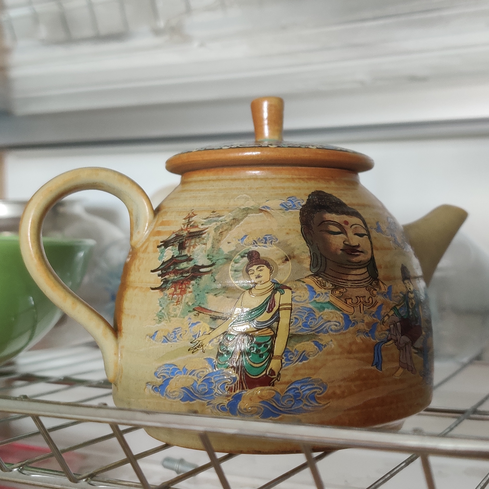 A ceramic teapot decorated with Buddhist imagery sits on a metal rack. The teapot has a light brown glaze with painted scenes of figures, pagodas, and clouds in shades of blue, green, and red. A handle extends from the side, and a spout is visible on the right. The background is out of focus, showing a white surface with horizontal lines, possibly a shelf or wall, and a blurred green object to the left. The teapot is positioned in a way that suggests it might be in a kitchen or display area. The image does not contain information that would allow for its location to be determined within Yekaterinburg, Russia.