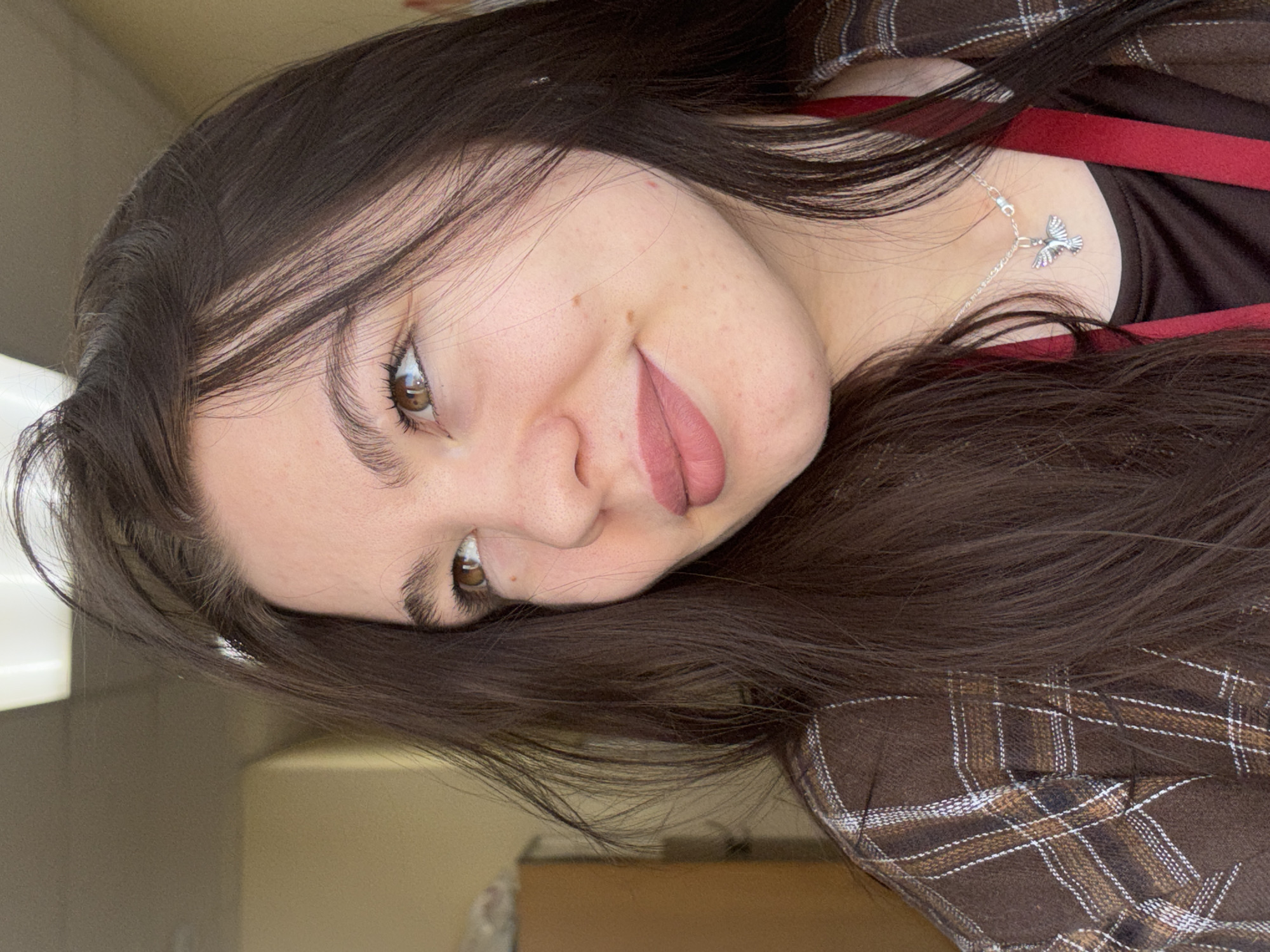 A young woman with long, dark brown hair and brown eyes is posing for a selfie. She is wearing a brown plaid shirt over a dark brown top with a red strap visible. She is also wearing a silver necklace with a bird pendant. Her lips are colored with a muted rose shade. The background is blurry and indistinct but appears to be an indoor space. There is no information to indicate the location in the city of Perm, Russia.