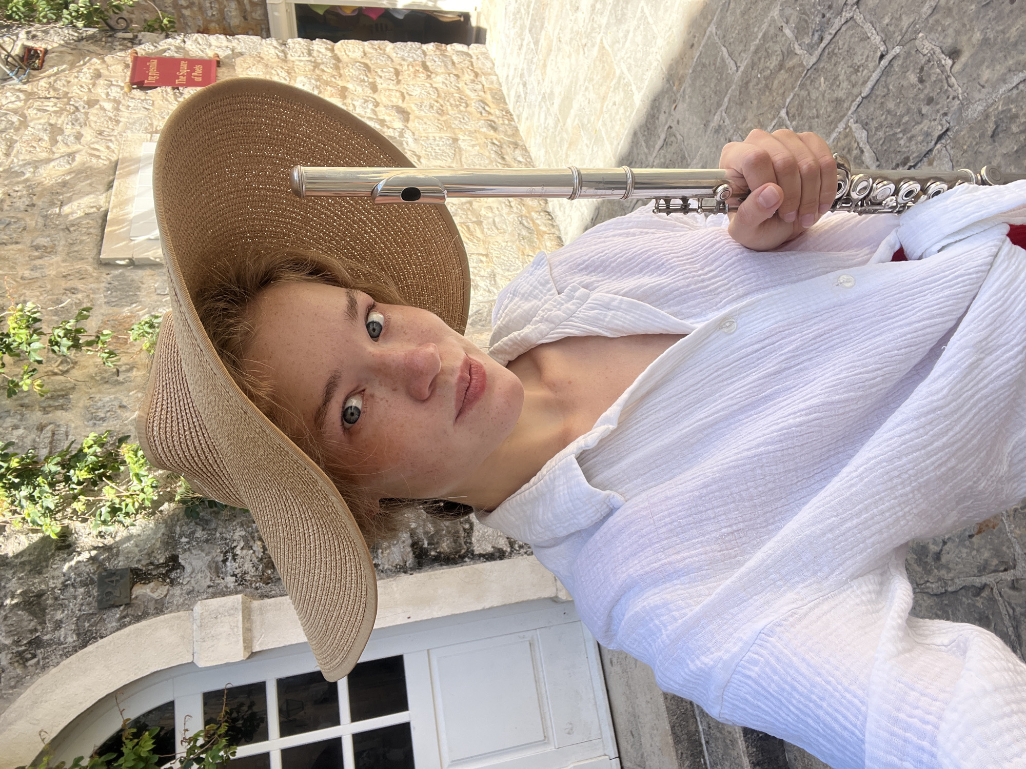 A fair-skinned, red-haired woman wearing a tan sun hat and a white button-up shirt is holding a silver flute while posing for a photo in the city of Budva, Montenegro. The woman is looking directly at the camera. Her skin has freckles. The background includes a building with a red sign indicating the "Square of Poets," as well as a white door with a grid of windows. The building is made of light-colored stone and is partially covered in greenery. The ground is paved with stone.