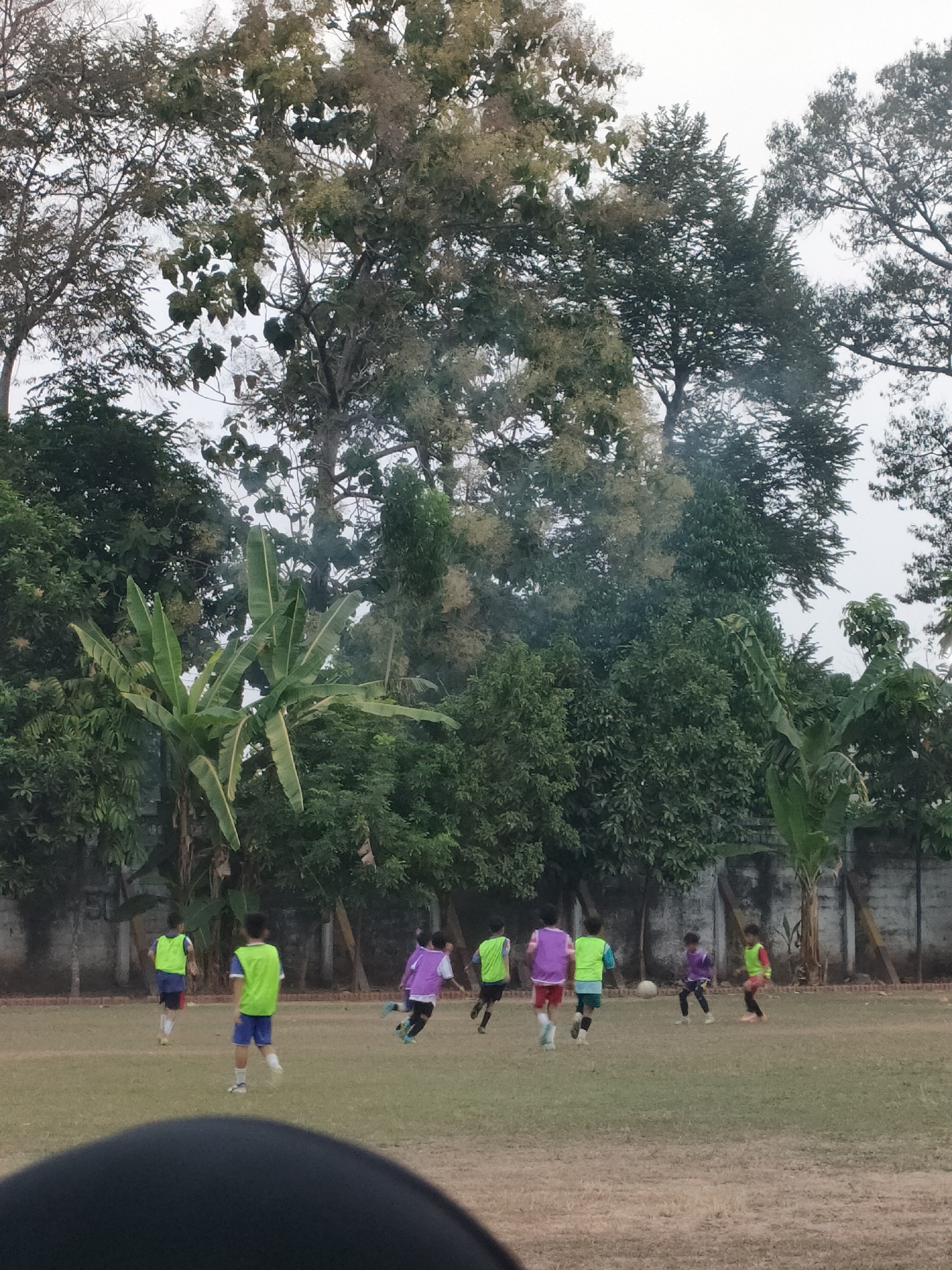The image depicts a group of children playing soccer on a field with a wall and trees in the background. The children are wearing colorful vests and shorts, and they are actively engaged in the game, running and kicking the ball. The field appears to be made of grass, and there are some trees and a wall bordering the playing area. The overall scene suggests a casual outdoor activity taking place in a somewhat rural or suburban setting, potentially within a park or a school ground. The presence of banana trees on the left side of the image might indicate a tropical or subtropical climate. The lighting suggests it could be late afternoon or early morning. This image is located in the city of Banguntapan, Indonesia.