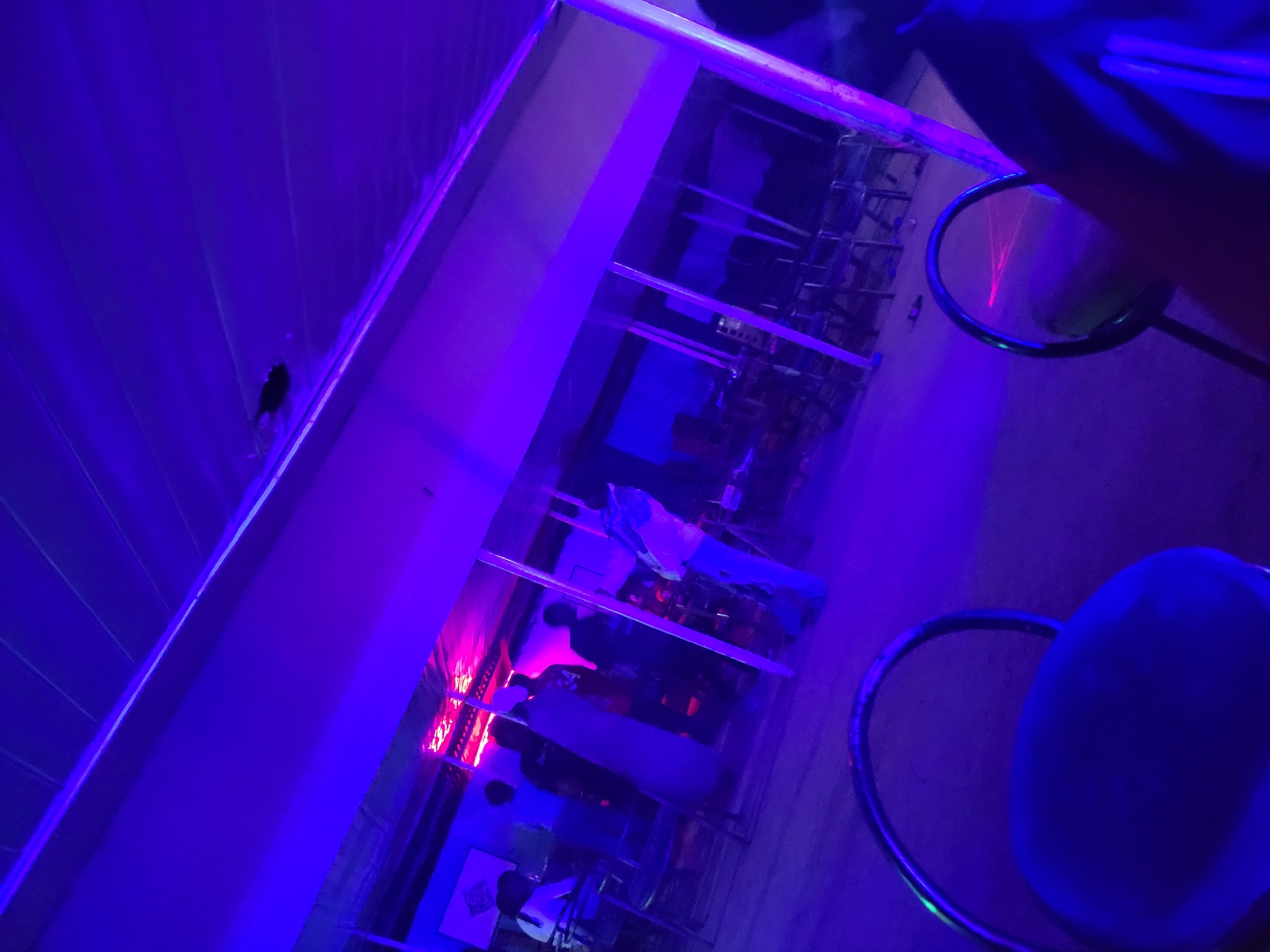 The image depicts an indoor venue, likely a bar or social gathering space, illuminated by vibrant blue and pink neon lights. Several individuals are visible, some standing and engaged in conversation, while others appear to be seated. The environment suggests a lively atmosphere, possibly a party or a nighttime establishment. The furniture includes chairs and tables, typical of such venues. The presence of what appears to be a ceiling fan or light fixture is also noted, contributing to the overall ambient lighting. The location is identified as Agwala, Nigeria.