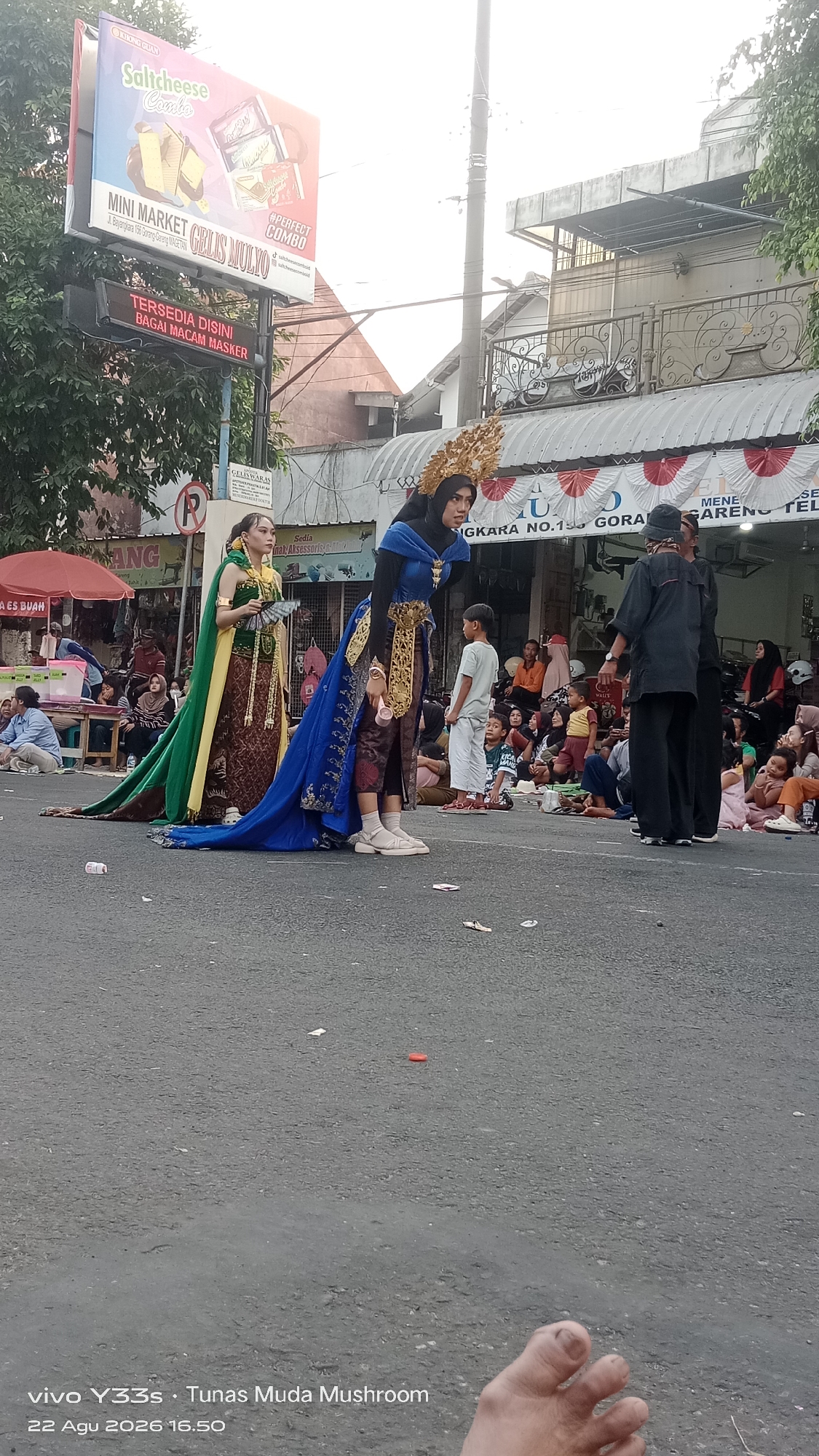 In the city of Genengan, Indonesia, on August 22, 2026, at 16:50, a public gathering is taking place on an asphalt street. Two individuals, adorned in elaborate traditional costumes, are the focal point of the scene. One person wears a vibrant blue gown with gold embellishments and a gold headdress, while the other is dressed in a green and gold outfit with a flowing cape, holding a fan. They appear to be part of a performance or cultural event.
A crowd of spectators, including adults and children, are seated on the ground, observing the performers. In the background, there are buildings with awnings and signage. One prominent billboard advertises "Saltcheese Combo" and "Mini Market Grilis Mulyo." Another sign below it states, "Tersedia Disini Berbagai Macam Masker" (Available Here Various Masks). Red and white banners are visible on the building awnings, suggesting a festive occasion. Several trees line the street, providing some shade. Debris, including a red plastic lid and paper wrappers, is scattered on the asphalt. The image is framed by a close-up of a person's foot in the lower right corner. The camera appears to be positioned at street level.