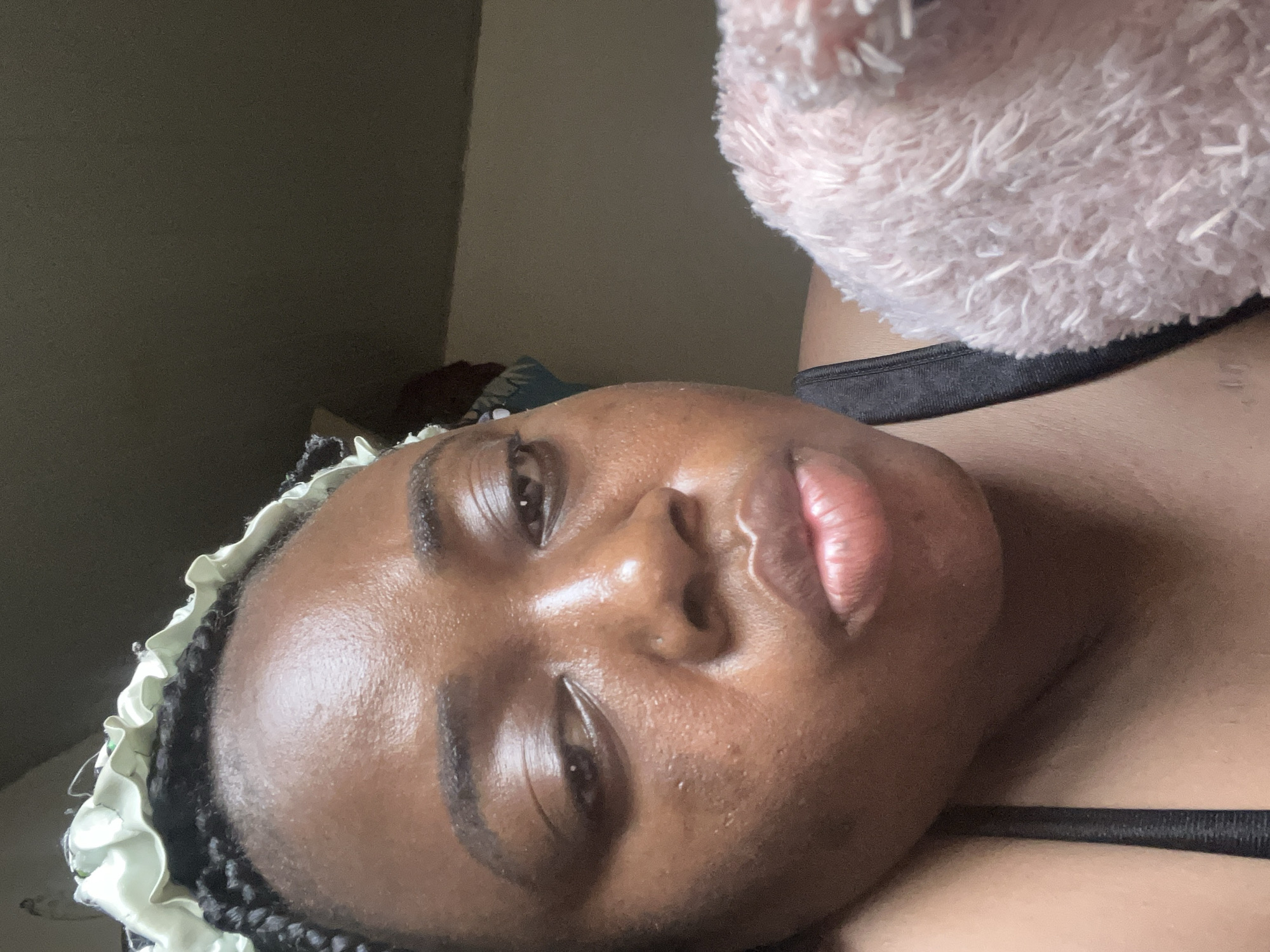 The image presents a close-up portrait of an individual, likely a young adult or adult female, positioned horizontally across the frame. She has dark skin, full lips with a natural pinkish hue, and dark eyes. Her expression is neutral to slightly pensive.
She is wearing a light-colored, possibly satin, ruffled bonnet or hair cover, under which dark braided hair is visible. A black bra strap or top is visible against her chest. In the upper right portion of the image, a soft, fluffy object of light pink color, resembling a blanket or plush item, is partially seen.
The subject is lying down, looking towards the camera. The background consists of a plain, dark-colored wall, suggesting an indoor setting, likely a bedroom in Abuja, Nigeria. The lighting is soft and ambient, casting subtle shadows but illuminating her features clearly. There is no visible text in the image.
