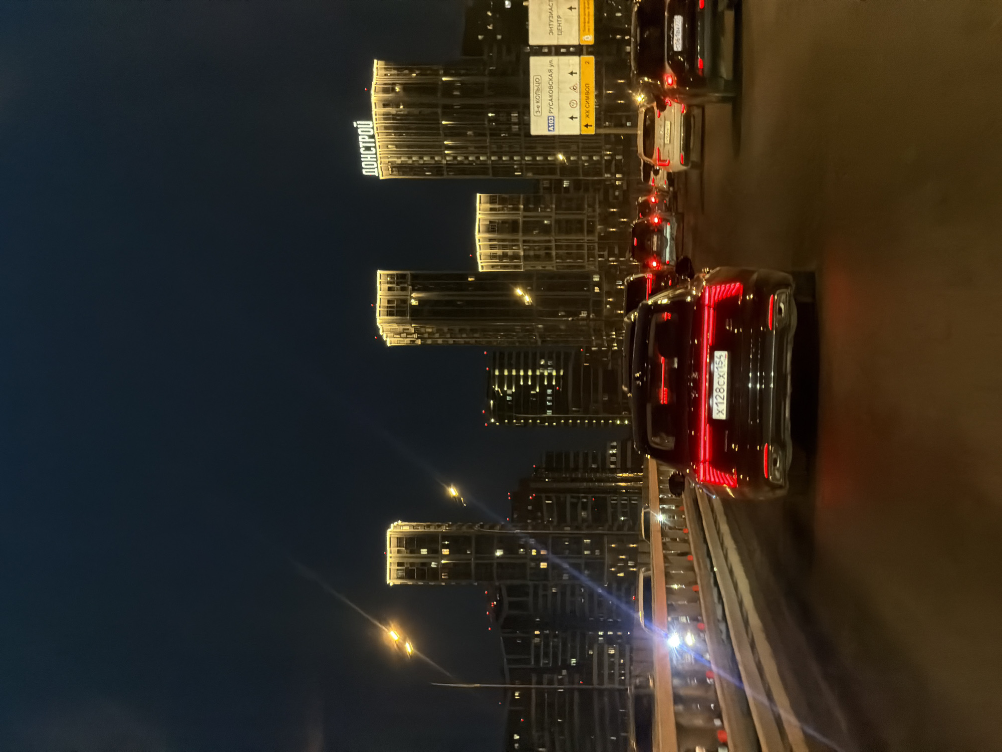 The image shows a nighttime view of Moscow, Russia, from the perspective of someone in traffic on a road. In the background are illuminated modern high-rise buildings, including one with the large word "ДОНСТРОЙ" (DONSTROY) lit up on top. A road sign indicates directions for "3-е кольцо" (Third Ring Road), "А103 РУСАКОВСКАЯ ул." (Rusakovskaya Street), "жк символ" (Symbol Residential Complex), "ЭНТУЗИАСТ" (Enthusiast) and "ЦЕНТР" (Center). The foreground features cars in traffic, with the tail lights of the nearest car prominently visible, including its license plate number "x128cx155". The streetlights are visible along with lights from the buildings in the distance.
