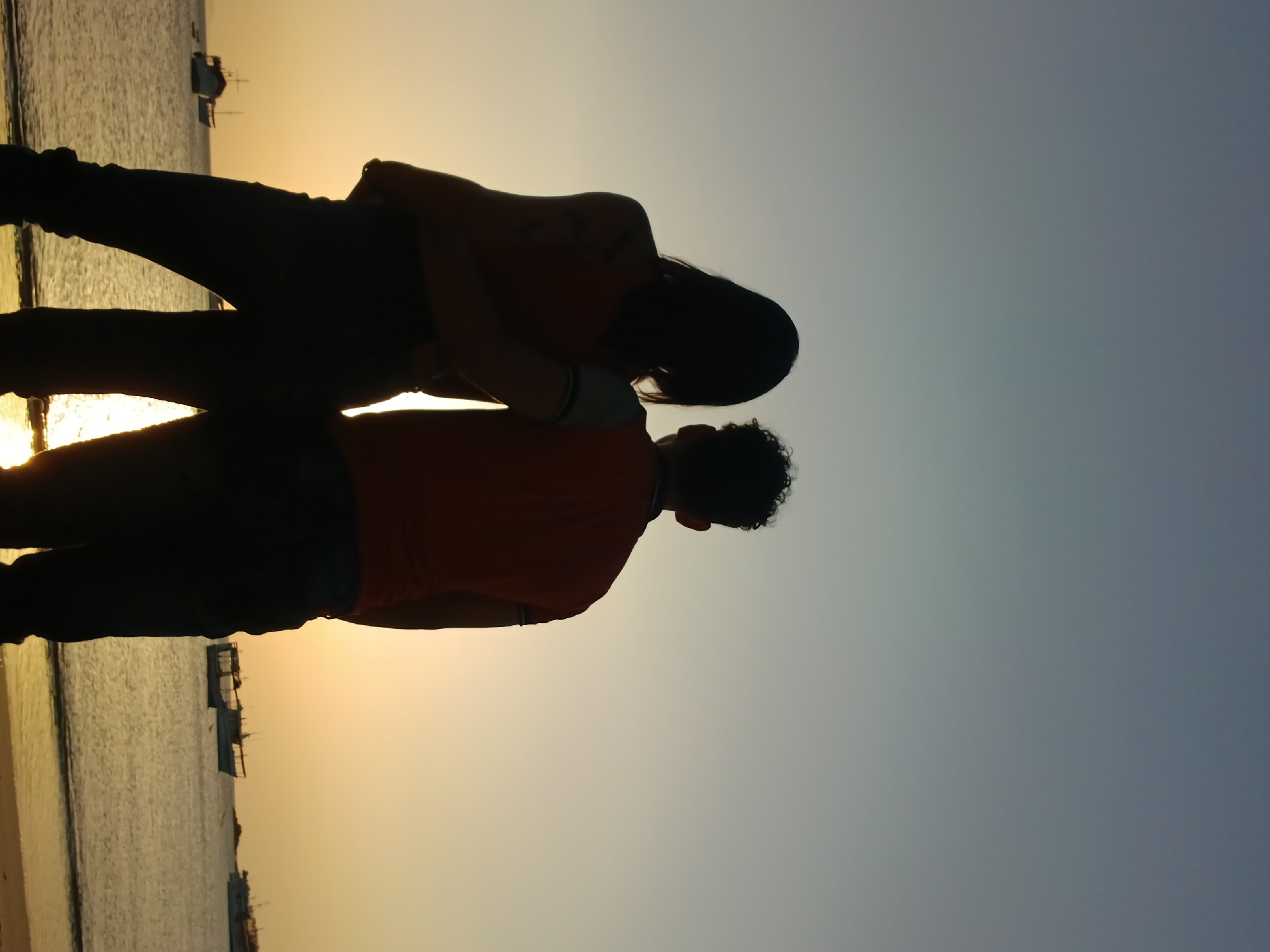 The image shows the silhouette of a couple embracing on a beach in Juan Griego, Venezuela, at sunset. The beach is sandy, and the water is visible in the background with a few boats on the horizon. The couple is wearing dark pants and lighter-colored shirts. The man has curly hair and is slightly shorter than the woman, who has long hair.