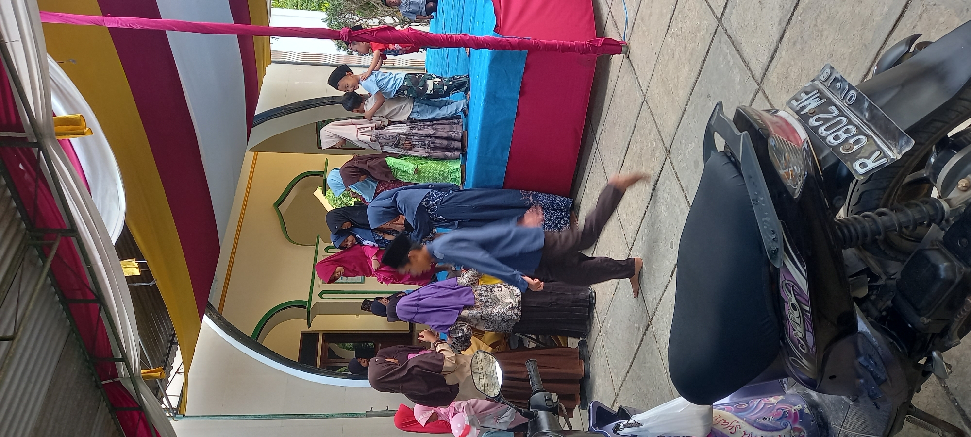 In Cintaratu, Indonesia, a group of children, dressed in traditional Islamic attire, stand on a raised blue platform decorated with red fabric. Some children are looking towards the camera, while others interact with each other. A young boy in a blue shirt and dark pants is captured mid-stride, running barefoot across the tiled ground in front of the platform. A motorcycle is parked in the foreground, its black seat and license plate clearly visible. The background features a building with an arched entrance, adorned with colorful fabric drapes overhead, suggesting a festive or ceremonial occasion. The overall scene depicts a lively community gathering.