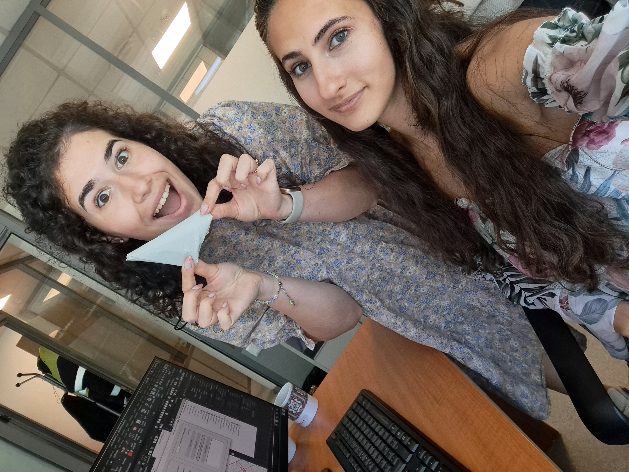Two young women are taking a selfie in an office environment. The woman in the foreground on the right has long, dark brown hair and is looking directly at the camera with a slight smile. She is wearing a floral-patterned top. The woman in the background on the left has curly dark brown hair and is holding a small piece of white paper, with a surprised expression and an open mouth. She is wearing a patterned dress. Behind the women, a computer monitor, keyboard, and a desk are visible. There is also a window with a view of what appears to be a hallway or another office space with coats hanging on a rack. The overall lighting suggests an indoor setting, likely during the daytime. The location is not identifiable from the image itself, and thus cannot be specified as Sofia, Bulgaria.