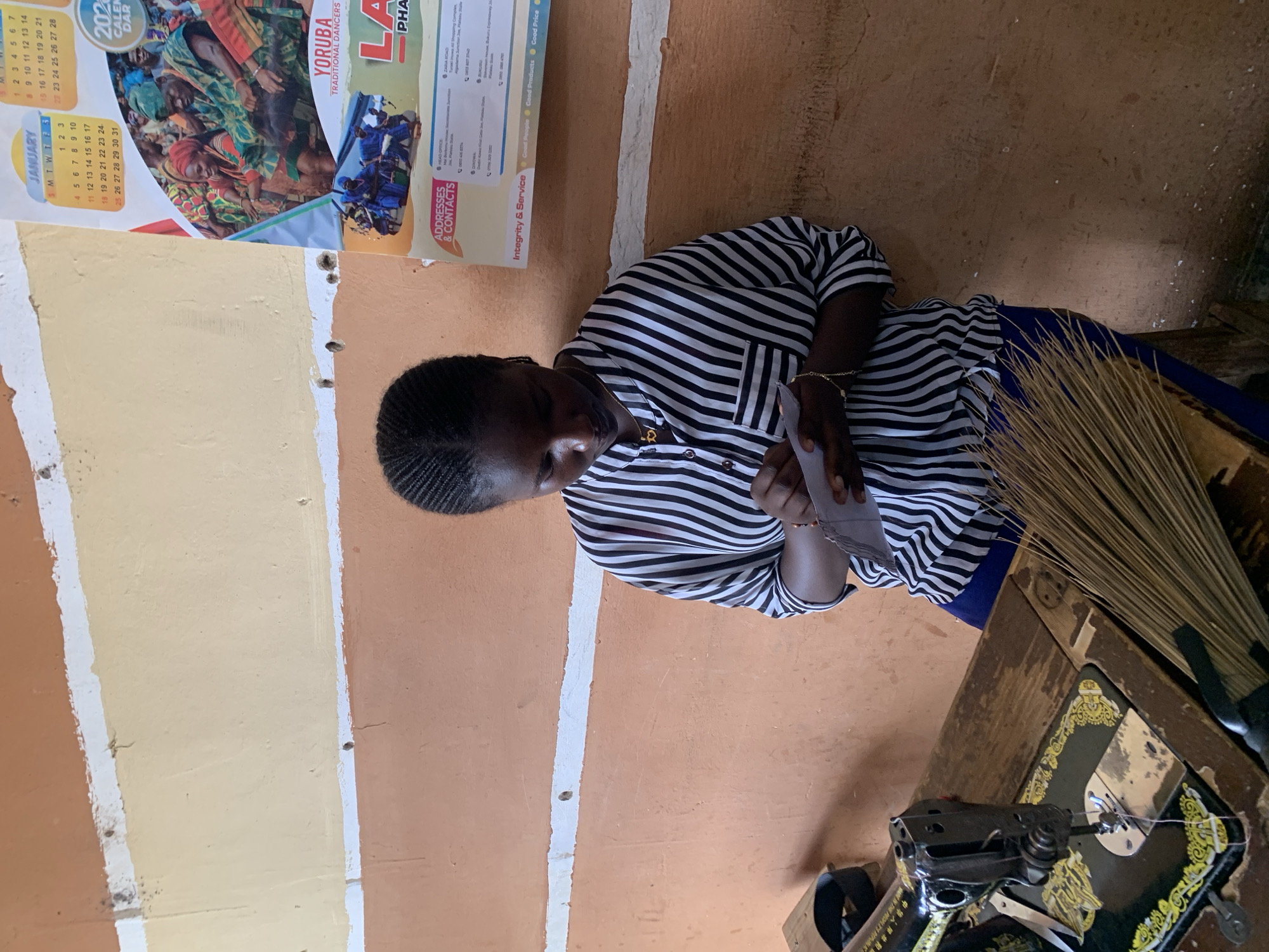 A woman sits at a sewing machine in a room with painted walls in bands of tan and beige. The room has a calendar showing January 2023 hanging on the wall above the woman, along with an advertisement for a company located in Jos, Nigeria. A broom leans against the wall near the sewing machine. The woman wears a black and white striped shirt and is looking down, holding paper in her hands.