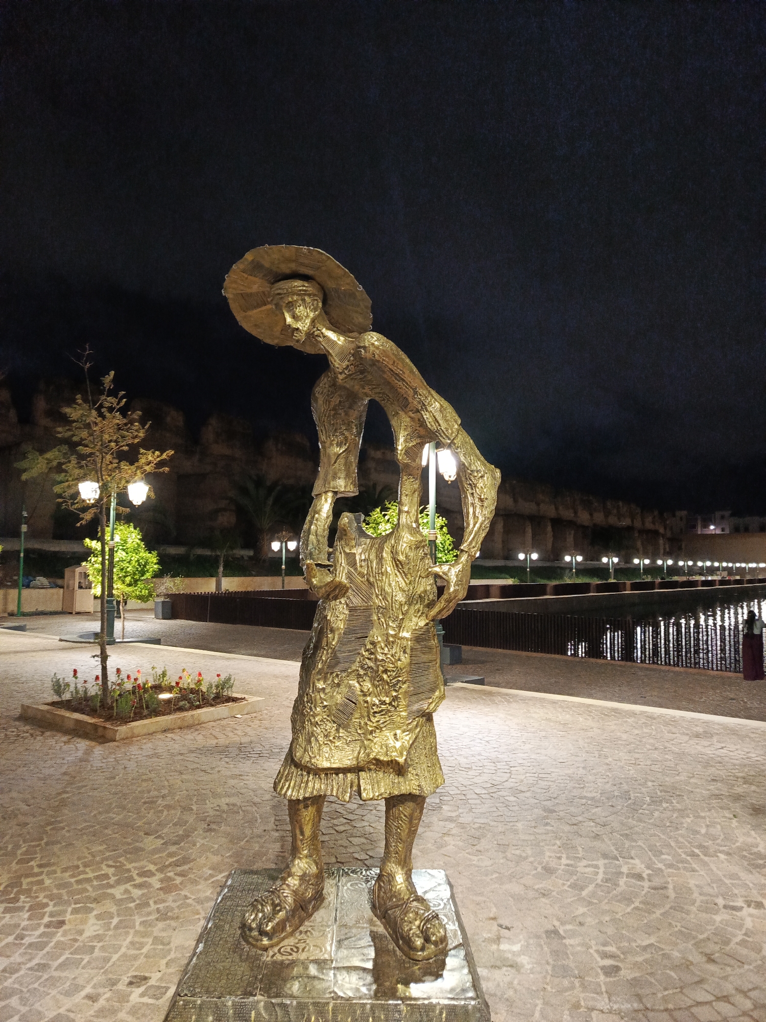 The image is a nighttime shot of a golden statue in the city of Al Machouar – Stinia, Morocco. The statue, depicting a figure with a wide-brimmed hat and wearing a dress or robe, stands on a stone-paved walkway. In front of the statue is a pool of water. Behind the statue are city walls, trees, and lampposts.
