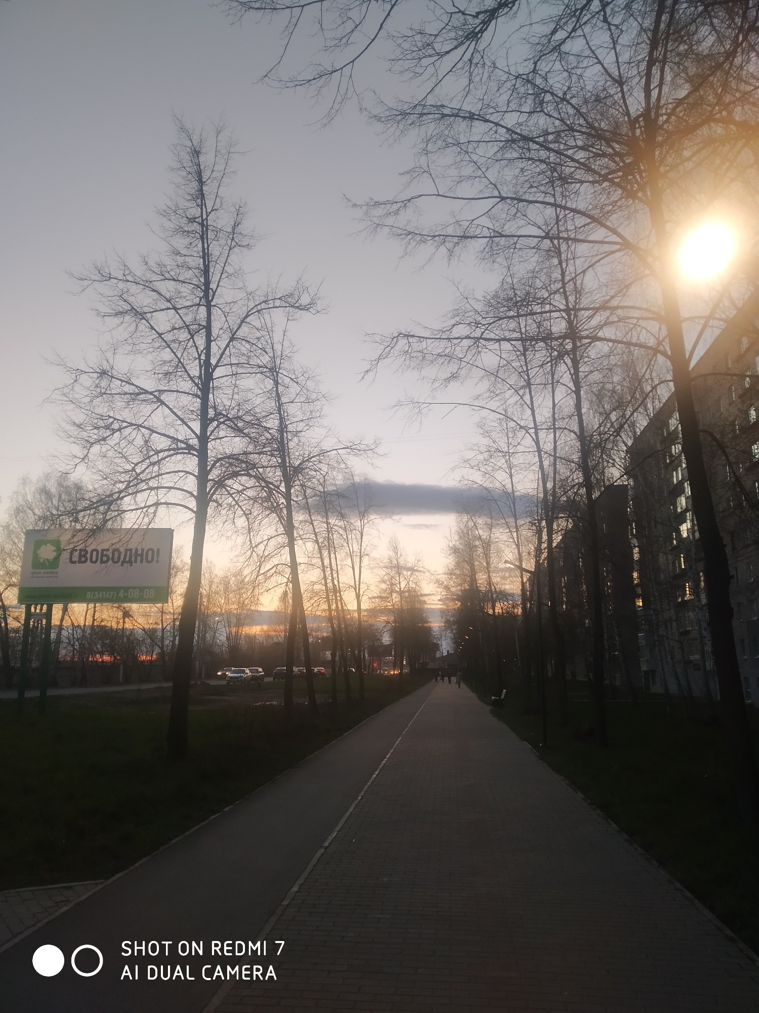 The image depicts an urban or suburban park pathway at dusk in Chaikovsky, Russia. A long, paved pedestrian path, possibly with a separate cycling lane, extends into the distance, flanked by green grass. Bare-branched deciduous trees line both sides of the path, silhouetted against the evening sky.
On the right, a large, multi-story residential building stands, with some windows showing internal lights or reflections of the fading sun. A bright streetlamp in the upper right casts a prominent glow. The sky transitions from a soft grey-blue directly overhead to warm orange and pink tones near the horizon, characteristic of a sunset.
On the left, a large billboard is prominently displayed. It features Cyrillic text that reads "СВОБОДНО!" (meaning "AVAILABLE!" or "FREE!"), along with "грин медиа" (green media), and a phone number: "8(34147)4-08-08." A small oak leaf logo is also visible on the billboard. Beyond it, several cars are parked or moving on a street. In the distance along the path, several indistinct figures appear as dark silhouettes, indicating people walking.
The scene suggests a quiet, routine evening in a residential area. A watermark in the bottom left corner reads "SHOT ON REDMI 7 AI DUAL CAMERA."