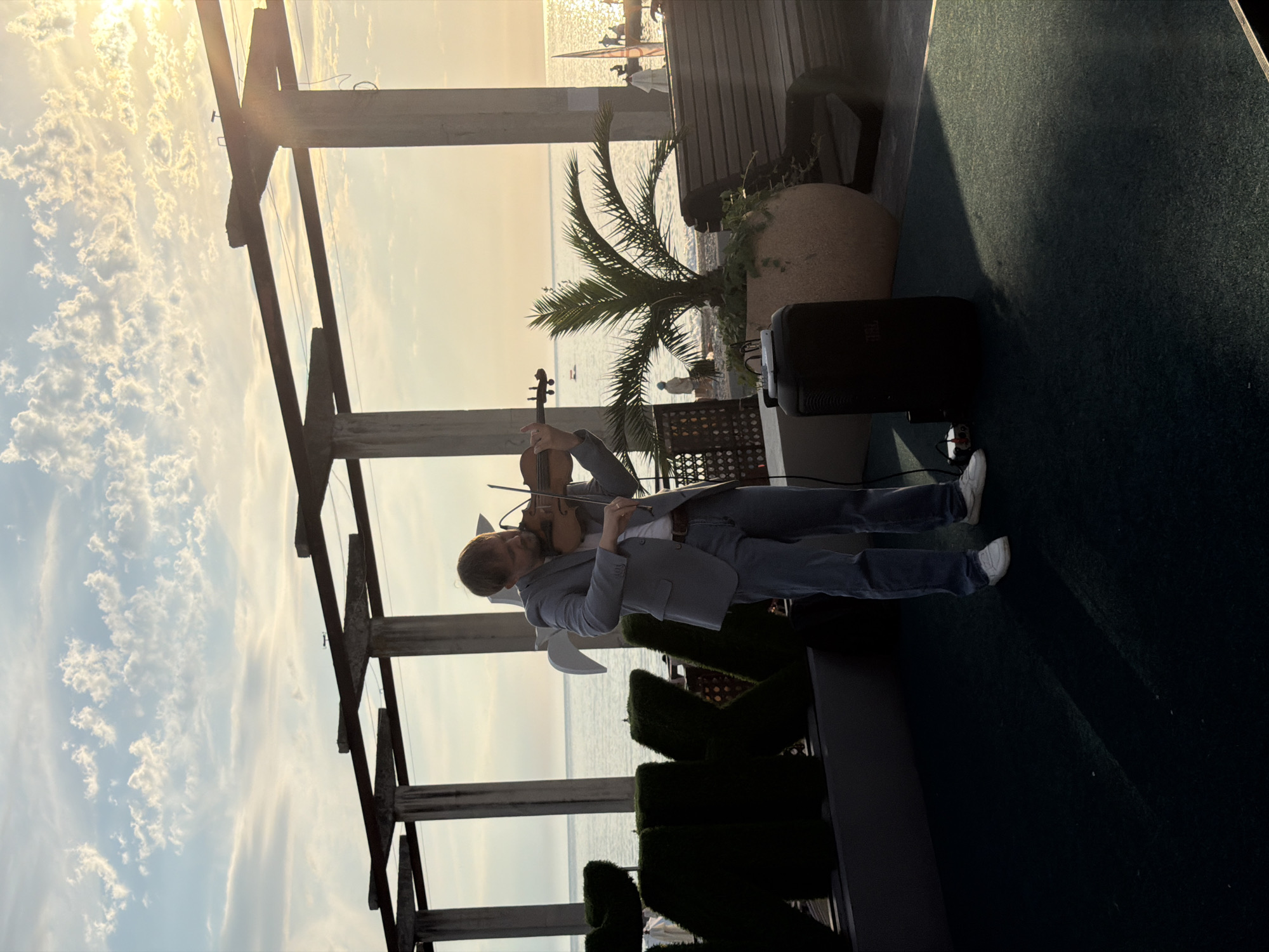 A man plays a violin on a stage near the sea in Sochi, Russia. The man is wearing a light blue suit jacket, a white t-shirt, blue jeans, and white sneakers. He is standing on a dark green carpeted stage with a black speaker to his right. Behind him, the sea is visible with boats and people in the distance. Above, a concrete structure with pillars and beams supports a roof, and a palm tree is planted in a large pot to the right of the man. The sky is blue with white clouds, and the sun is setting, casting a warm glow over the scene. Large green hedges shaped like letters are visible to the left of the stage.
