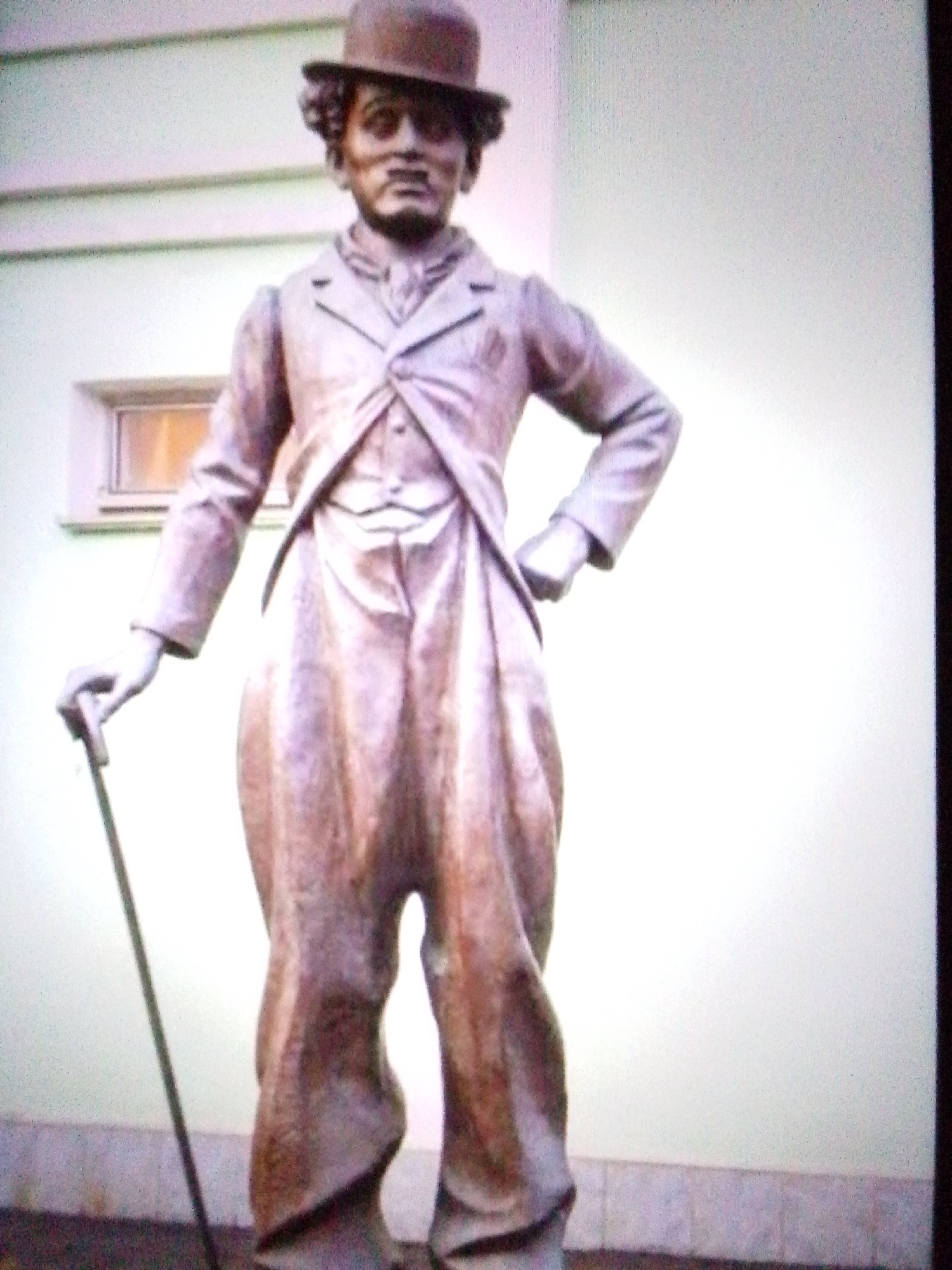 A full-body bronze statue of Charlie Chaplin as The Tramp stands outdoors in front of a light-colored building. The statue is detailed and appears to be made of weathered bronze. Chaplin is depicted wearing his signature bowler hat, suit, and oversized pants. He holds a cane in his left hand, which is resting on his hip. His right hand is out of view behind his back. The background consists of a building with a window and a light-colored wall. The ground beneath the statue appears to be paved. The overall lighting suggests it is daytime.